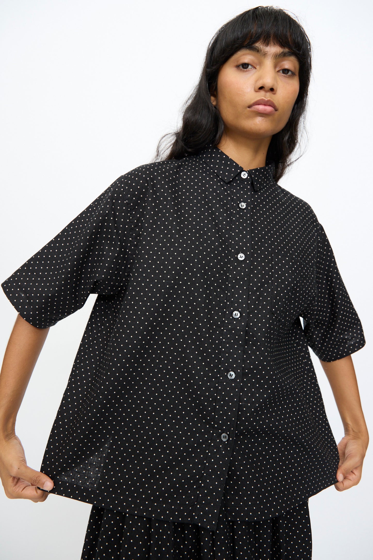 A person stands against a plain background, holding out an oversized Ichi Antiquités Cotton Linen Dot Shirt in Black— a loose-fitting, short-sleeve shirt with small white polka dots.