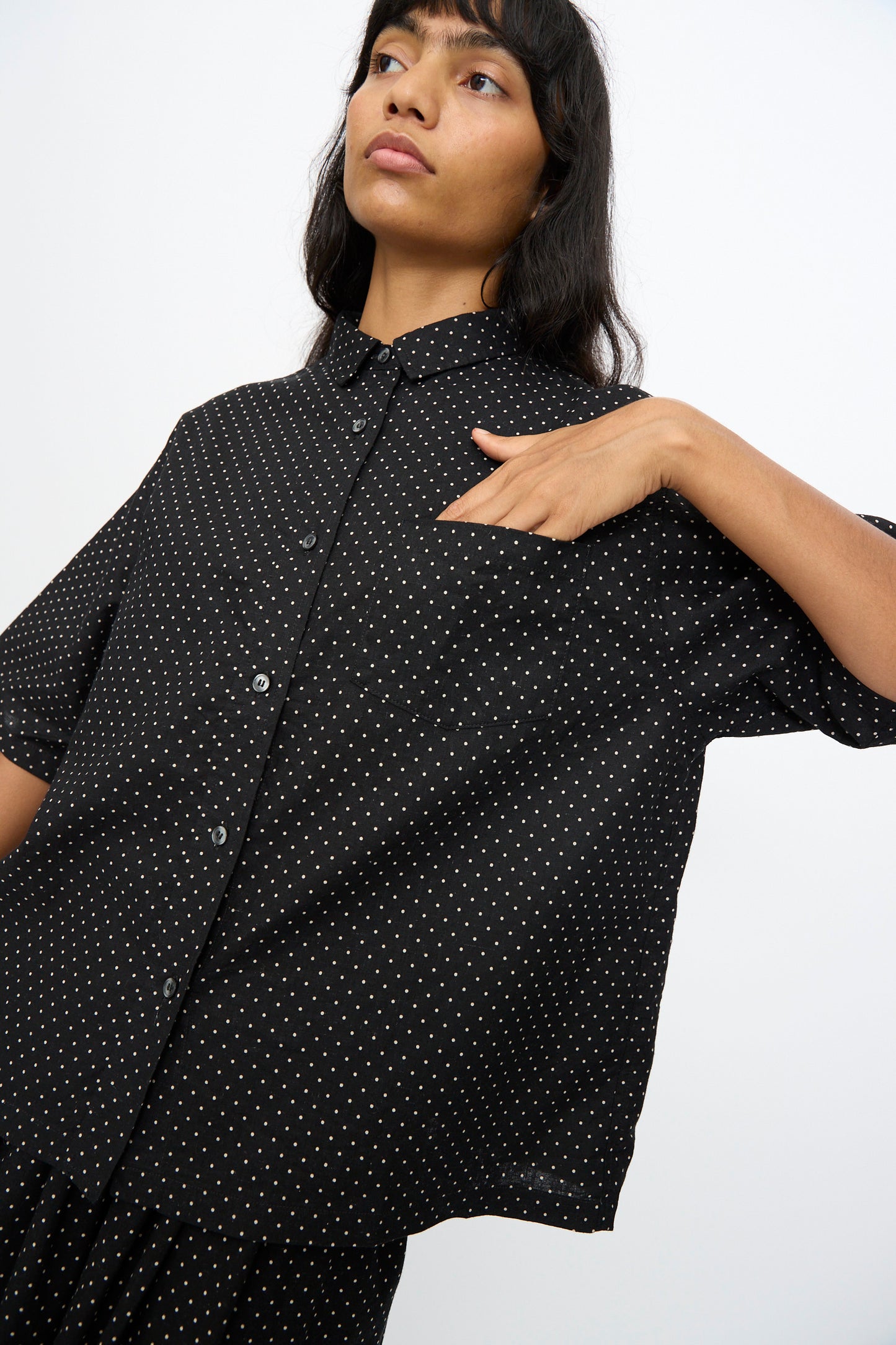A person wears the Ichi Antiquités Cotton Linen Dot Shirt in Black, an oversized short-sleeve shirt with polka dots, touching their chest against a plain white background.