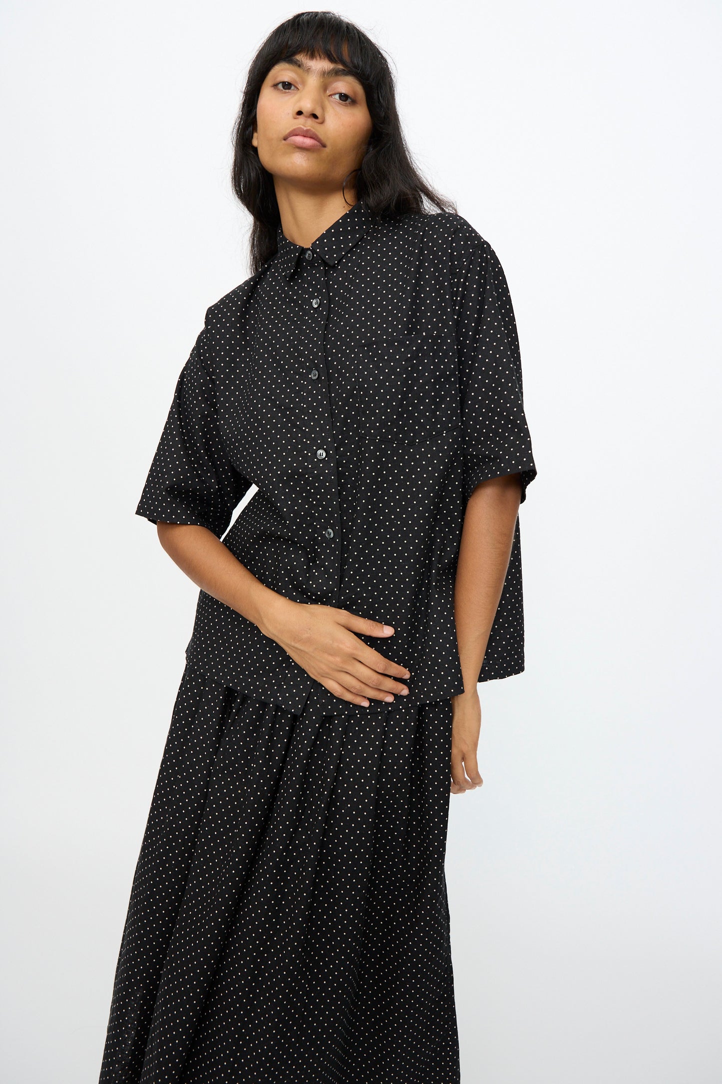 A woman stands against a plain light background wearing the Ichi Antiquités Cotton Linen Dot Shirt in Black, styled oversized with short sleeves, paired with a matching long skirt.