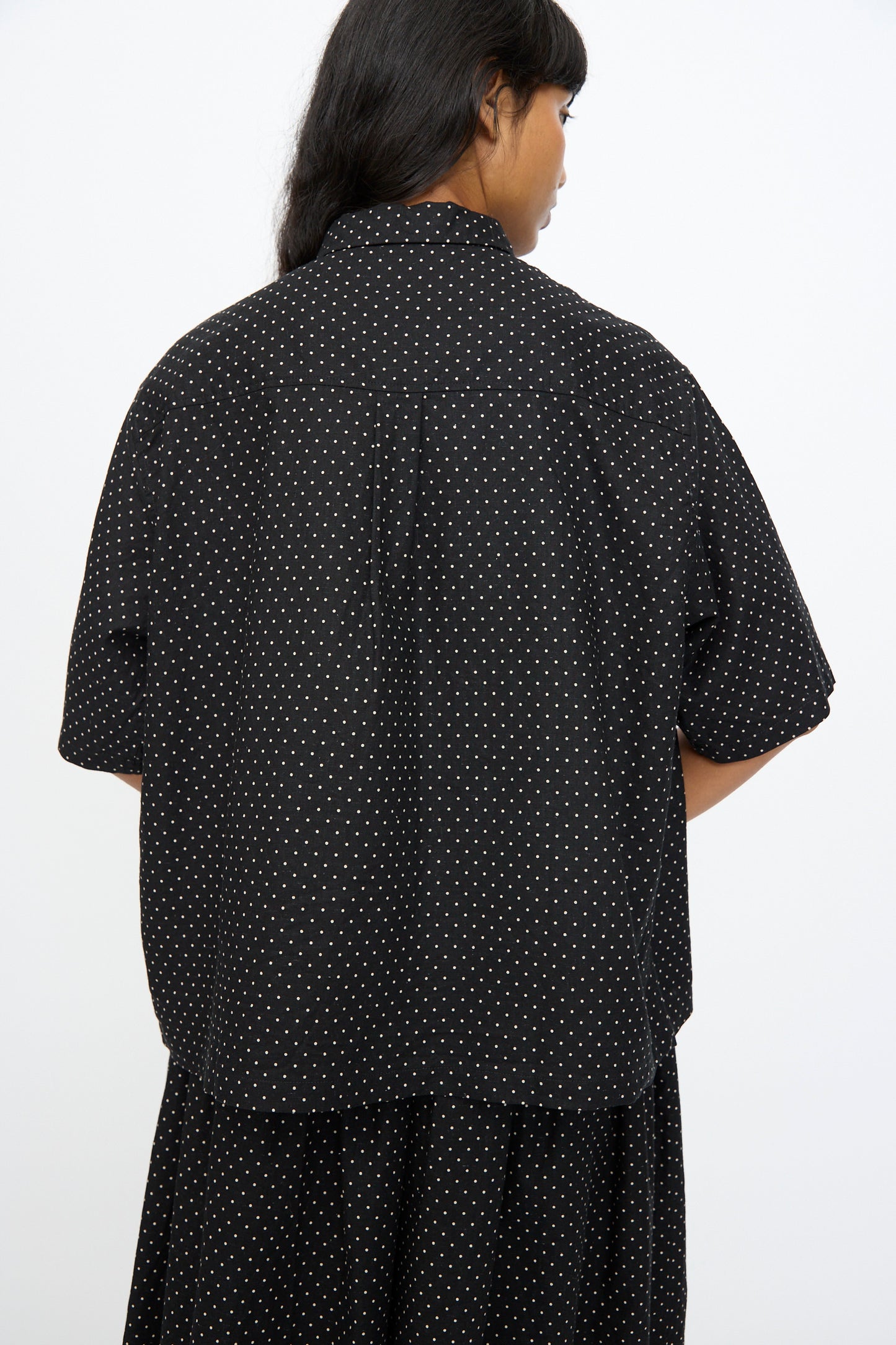 A person with long dark hair faces away, wearing the Ichi Antiquités Cotton Linen Dot Shirt in Black—an oversized shirt with small white dots—against a plain, light background.