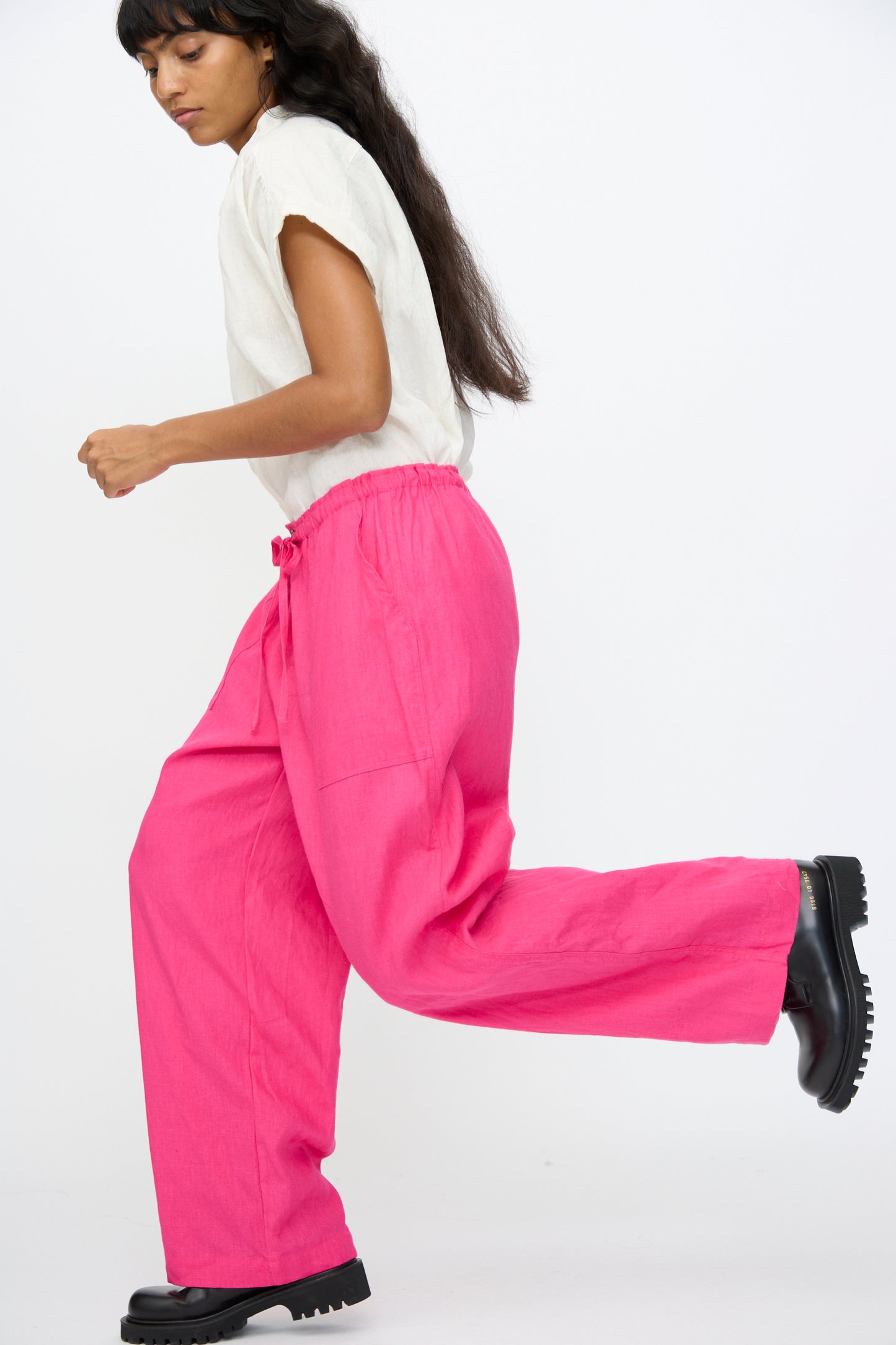 Someone is seen mid-step against a plain white background, wearing a short-sleeved white top, Ichi Antiquités Color Linen Pants in Pink with a relaxed fit, and black shoes.
