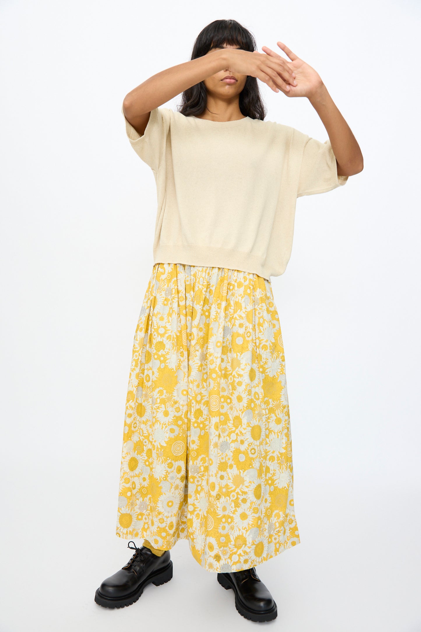 Wearing the Ichi Antiquités Cotton Linen Pullover in Natural, a yellow floral skirt, and black boots, a person stands with arms raised, partially covering their face against a plain white background.