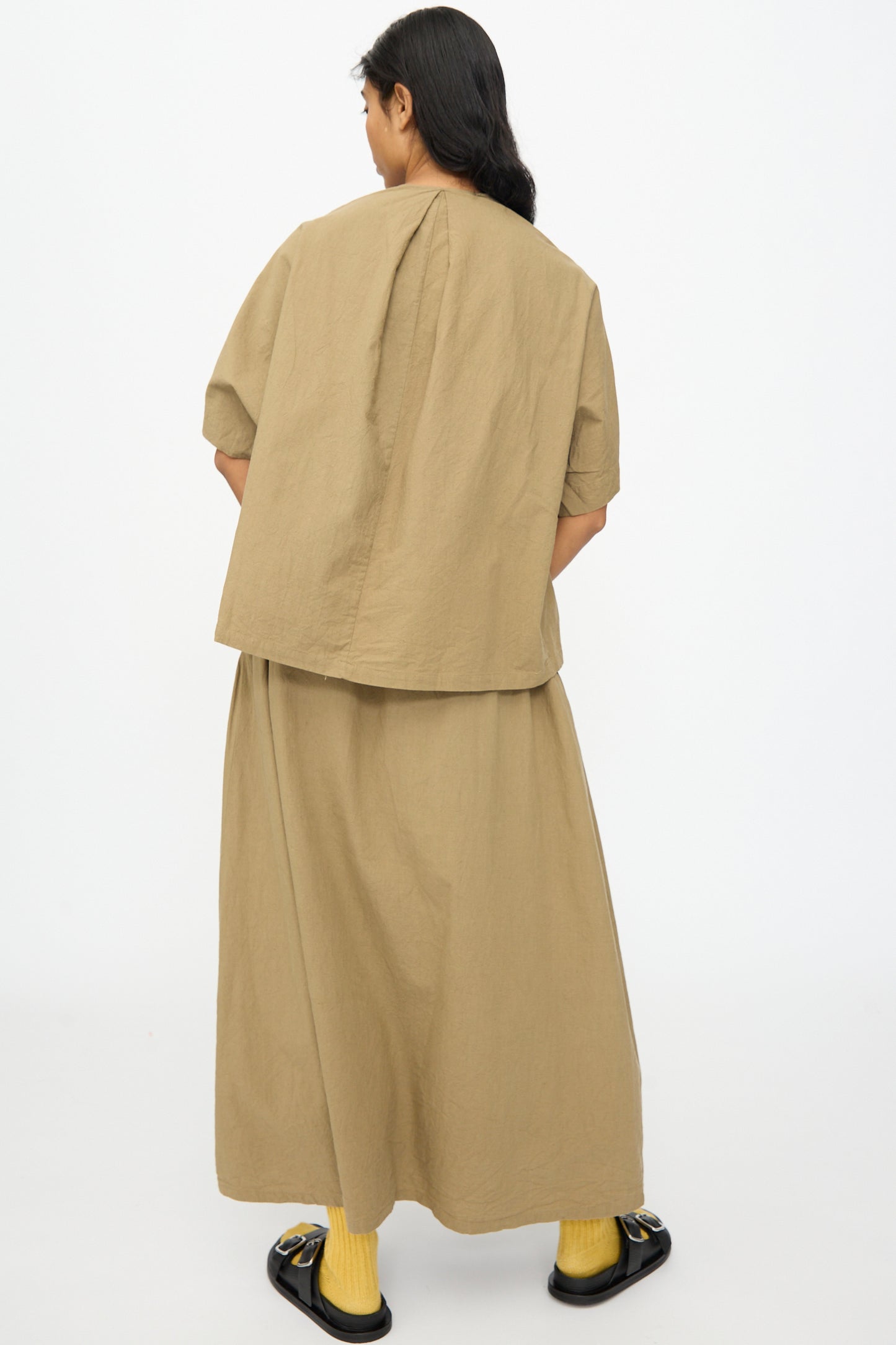 A person stands facing away against a plain white background, wearing the Ichi Antiquités Cotton Linen Shirt in Mocha with a matching long skirt, yellow socks, and black sandals.