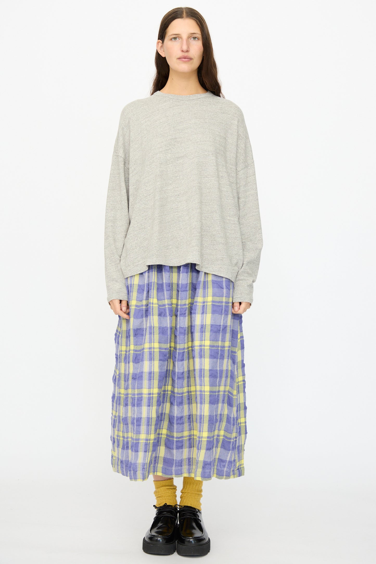 A person stands facing forward against a plain white background, wearing the Ichi Antiquités Cotton Loose Pullover in Gray, styled with a blue and yellow plaid midi skirt, mustard socks, and black shoes.