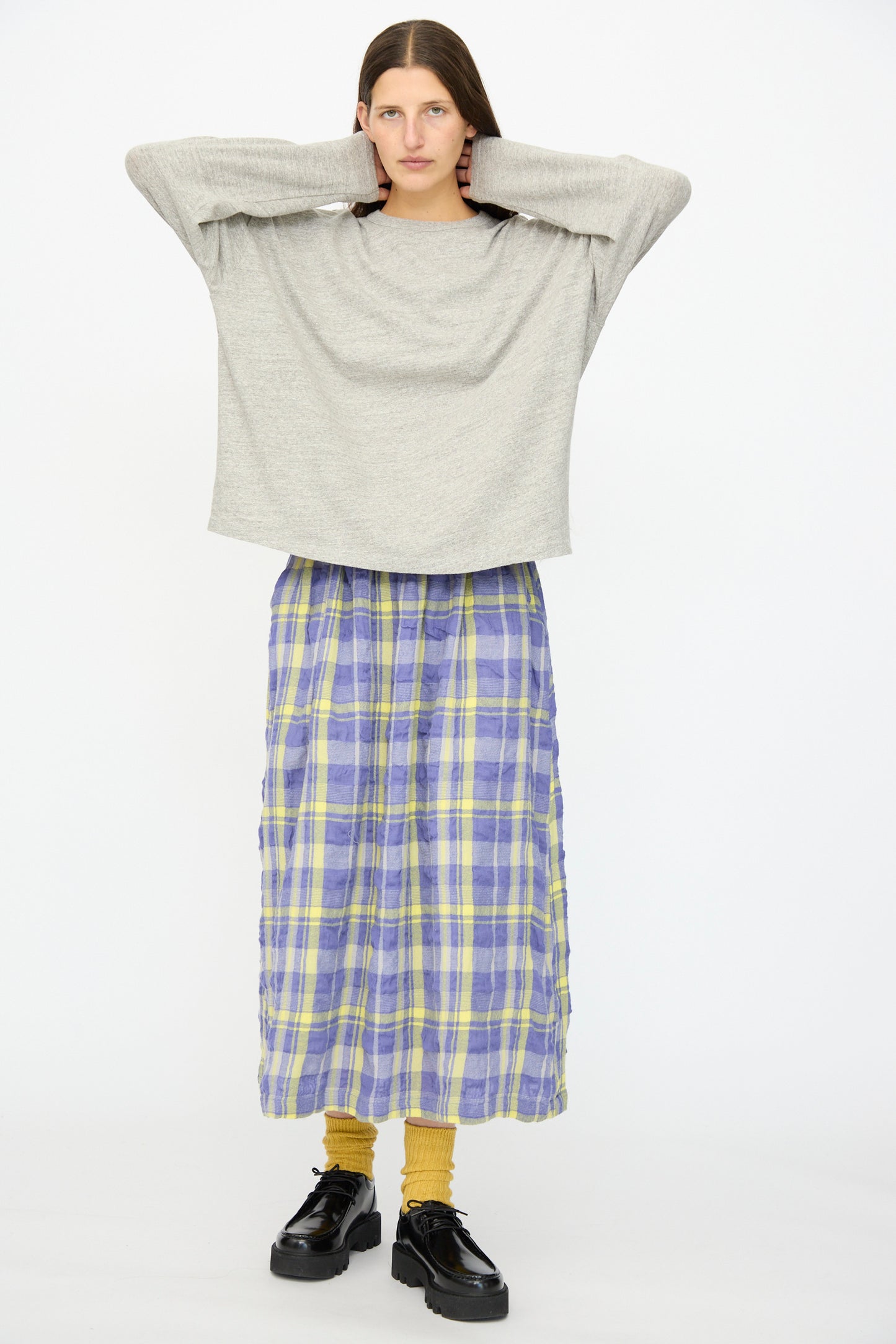 A person stands with hands behind head, wearing the Ichi Antiquités Cotton Loose Pullover in Gray, a long purple and yellow plaid skirt, mustard socks, and black platform shoes against a plain white background.