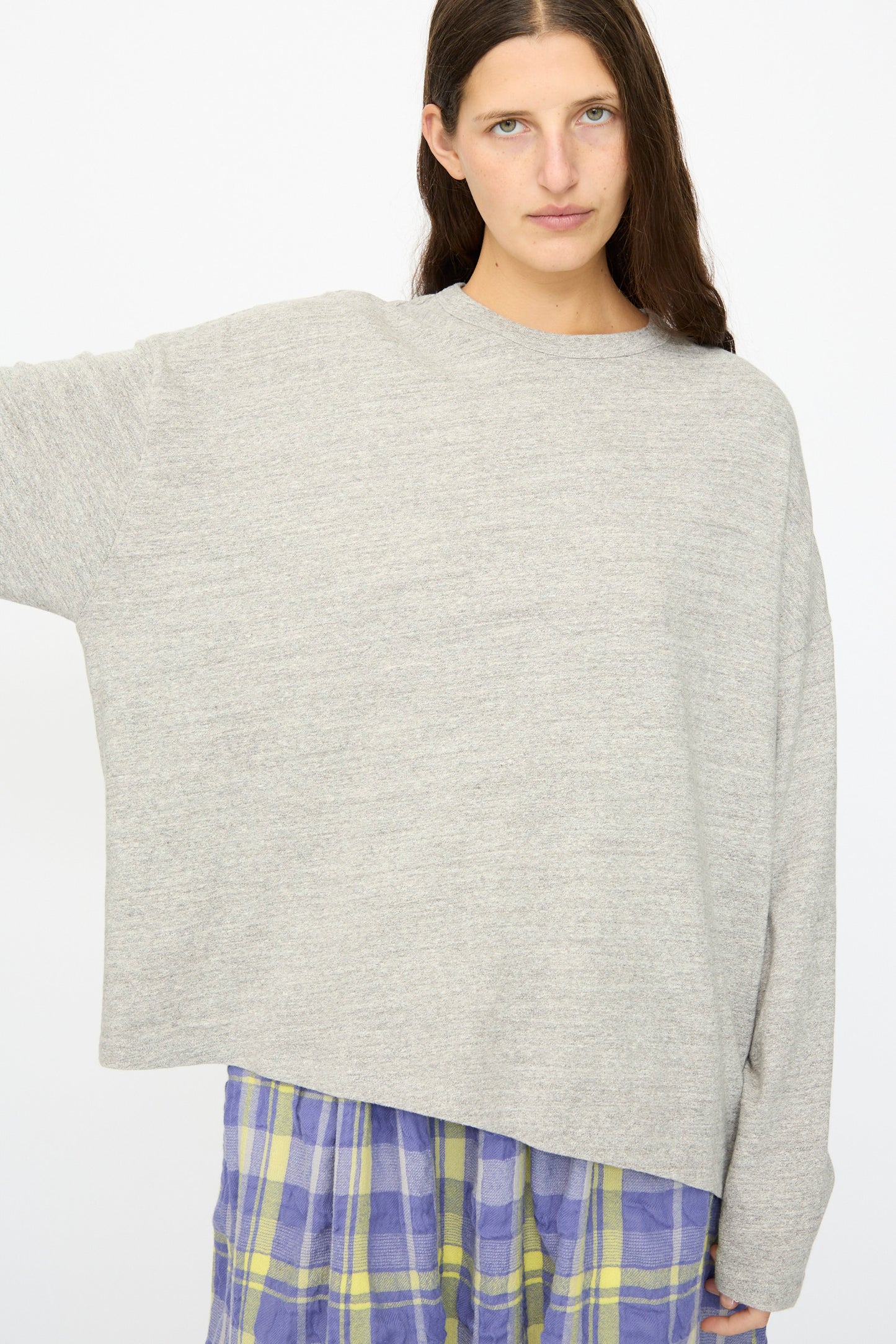 A person with long brown hair wears the Ichi Antiquités Cotton Loose Pullover in Gray and a blue and yellow plaid skirt, standing before a white background for an effortlessly cozy look.