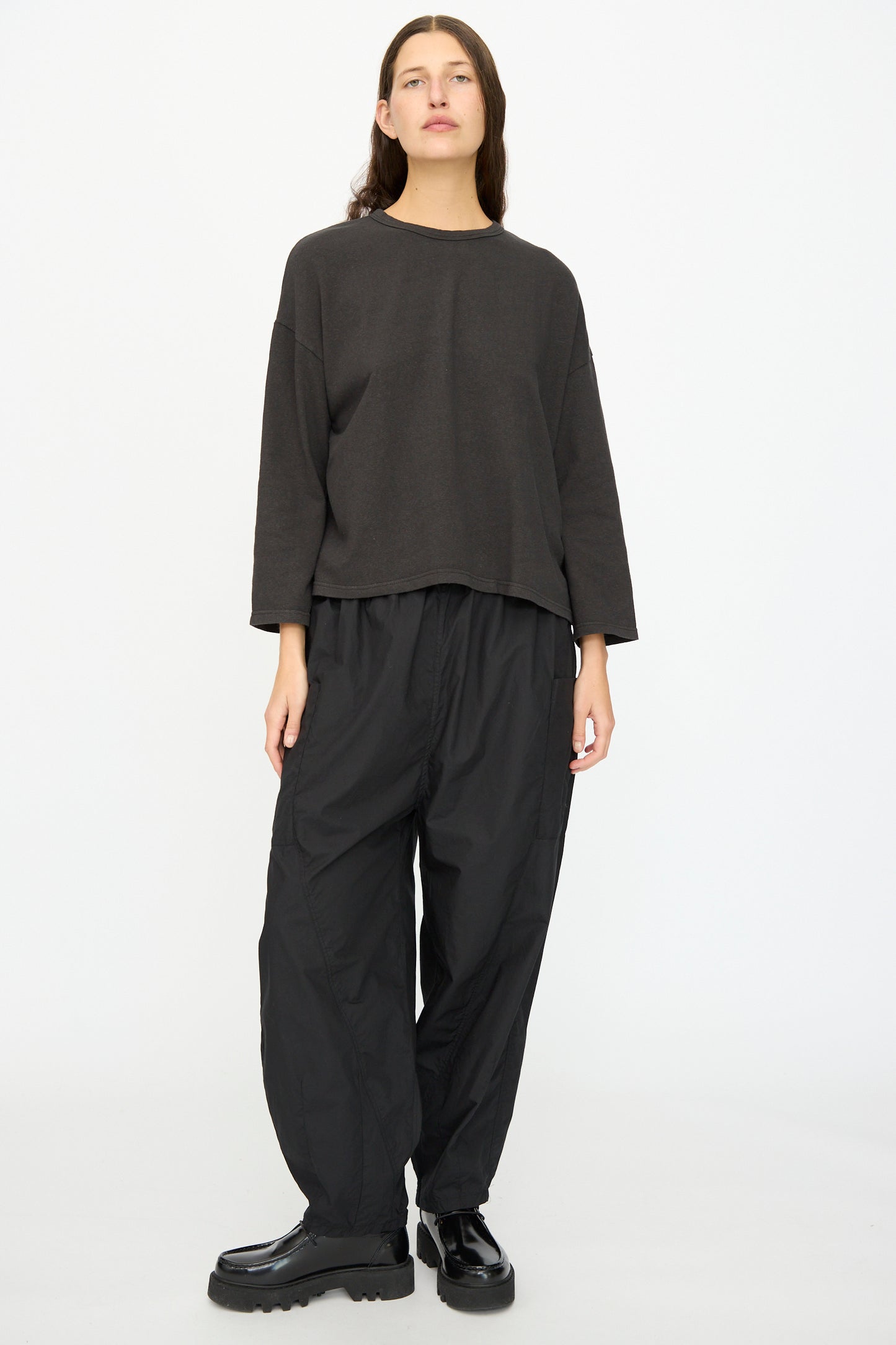 A person stands against a plain white background wearing the Ichi Antiquités Cotton Pullover in Black, paired with baggy black pants and black platform shoes.