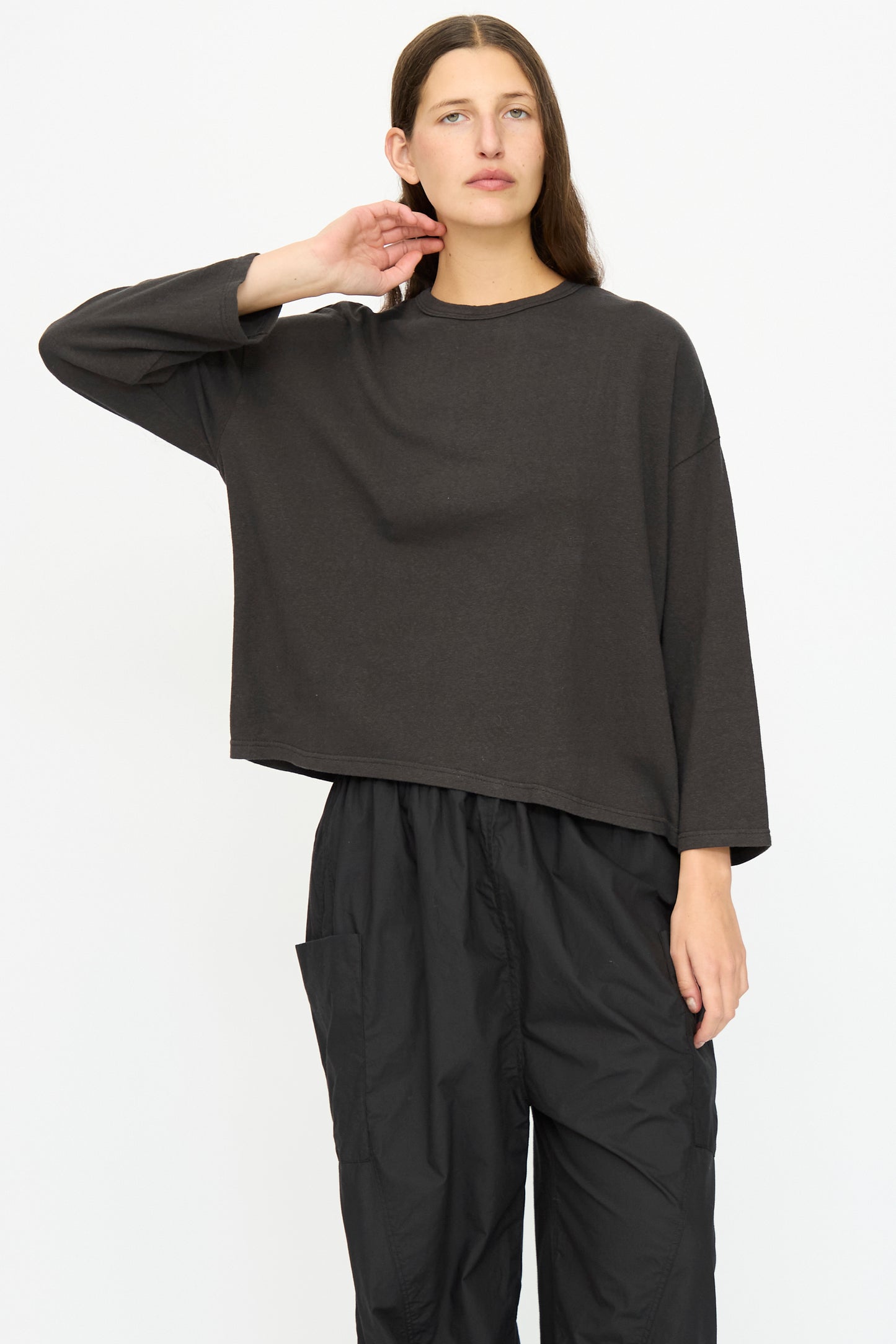 A person stands against a white background, wearing the Ichi Antiquités Cotton Pullover in Black—a relaxed fit top—paired with black pants featuring a pocket, with one hand touching their neck.
