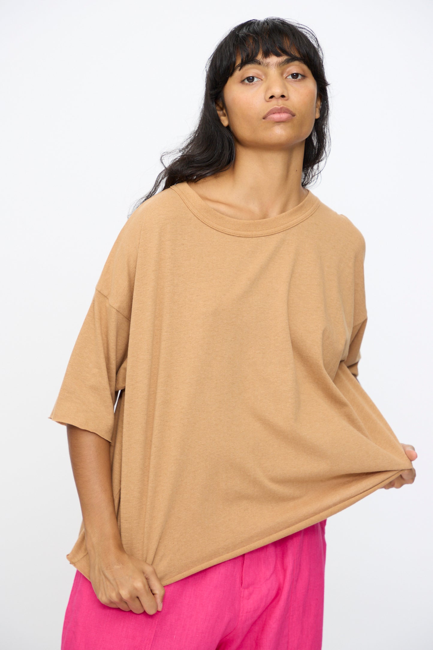 A person with long dark hair wears the Ichi Antiquités Cotton Pullover Tee in Camel, styled loosely, and bright pink pants, standing against a plain white background while gently pulling at the shirt's hem.