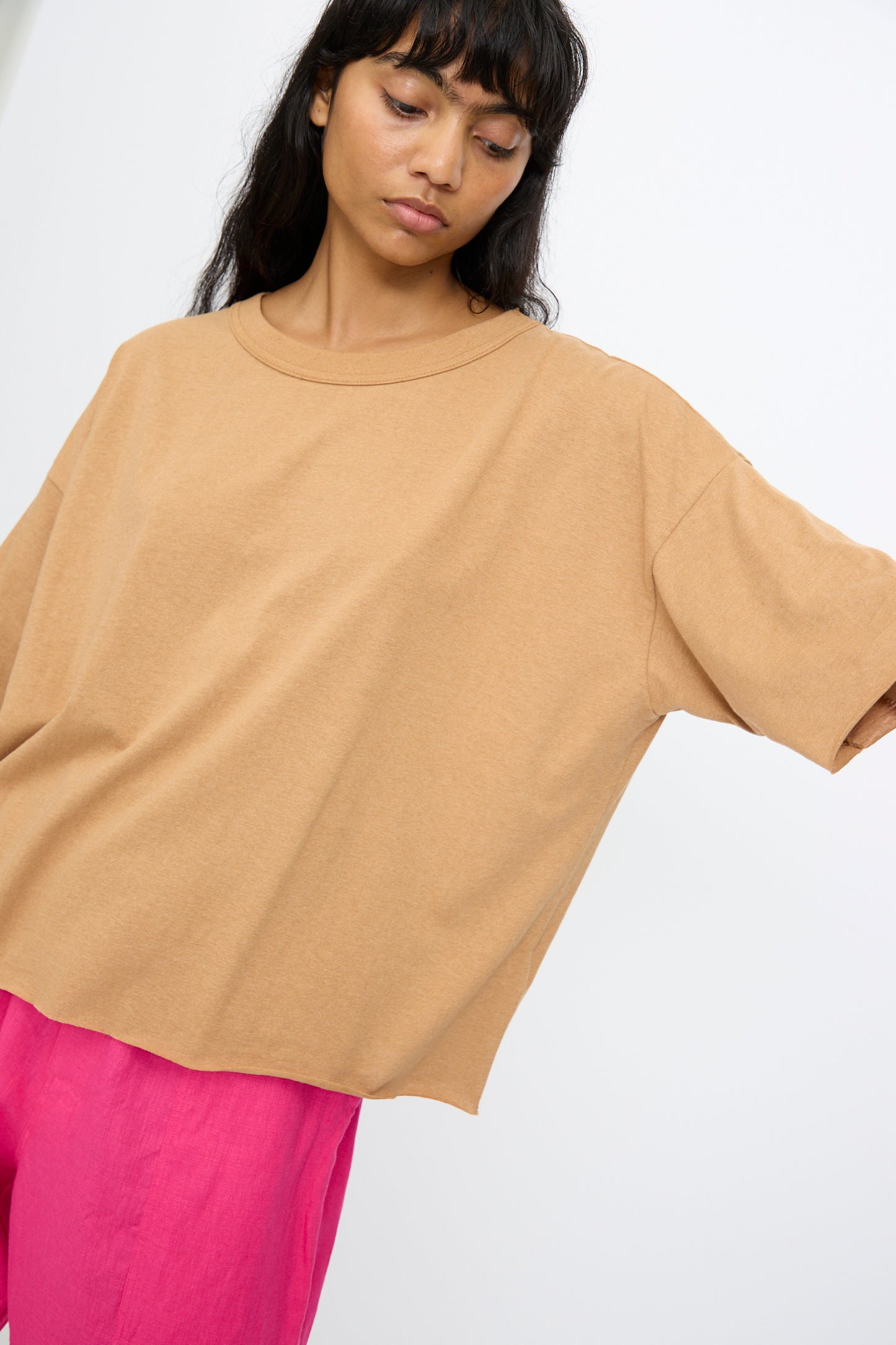 Someone wears the Ichi Antiquités Cotton Pullover Tee in Camel— a loose-fitting, made in Japan top—paired with bright pink pants, standing against a plain white background with one arm slightly raised.