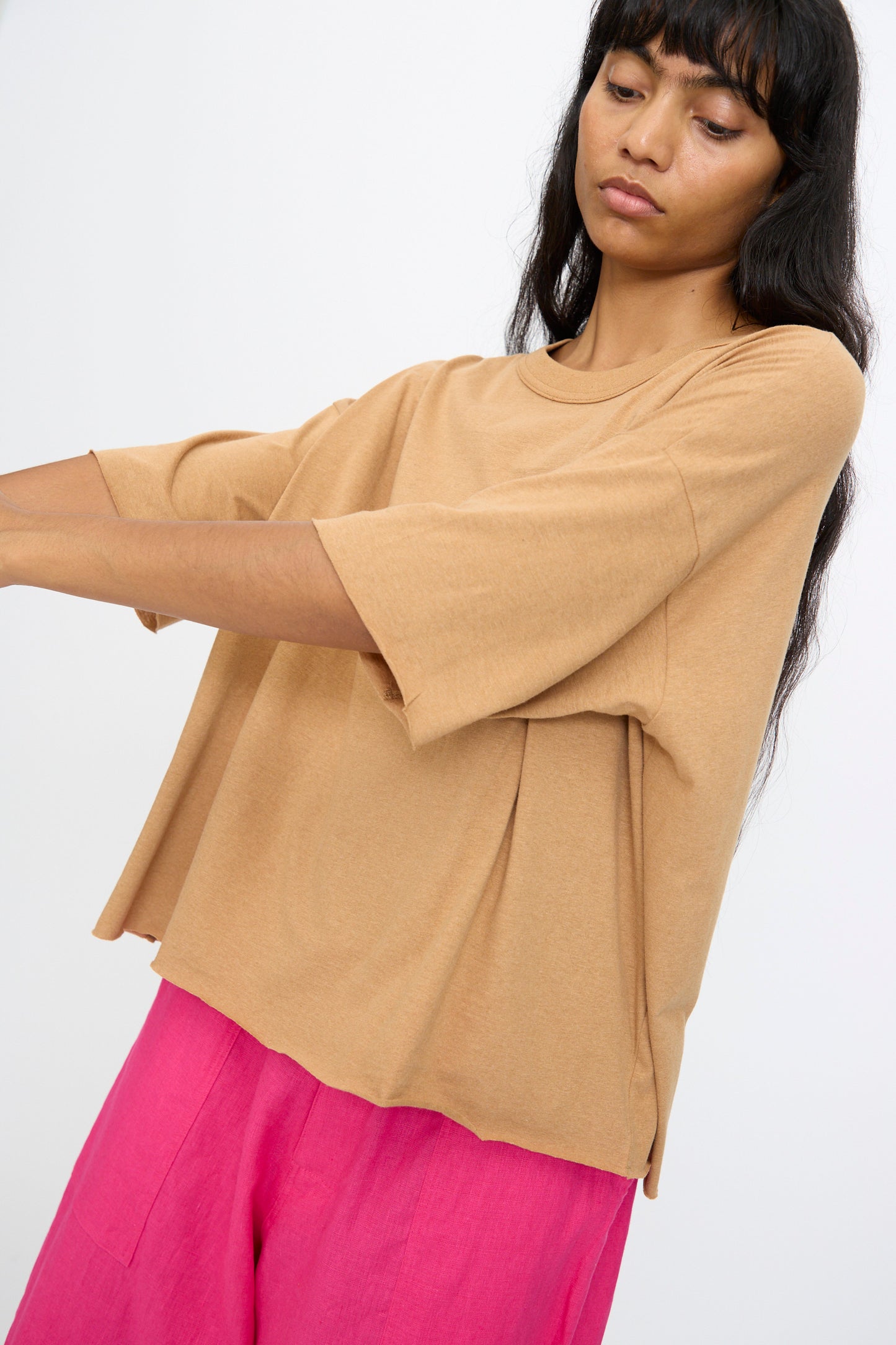 Model wears the Ichi Antiquités Cotton Pullover Tee in Camel, styled with bright pink pants and standing against a plain light background with arms slightly extended.