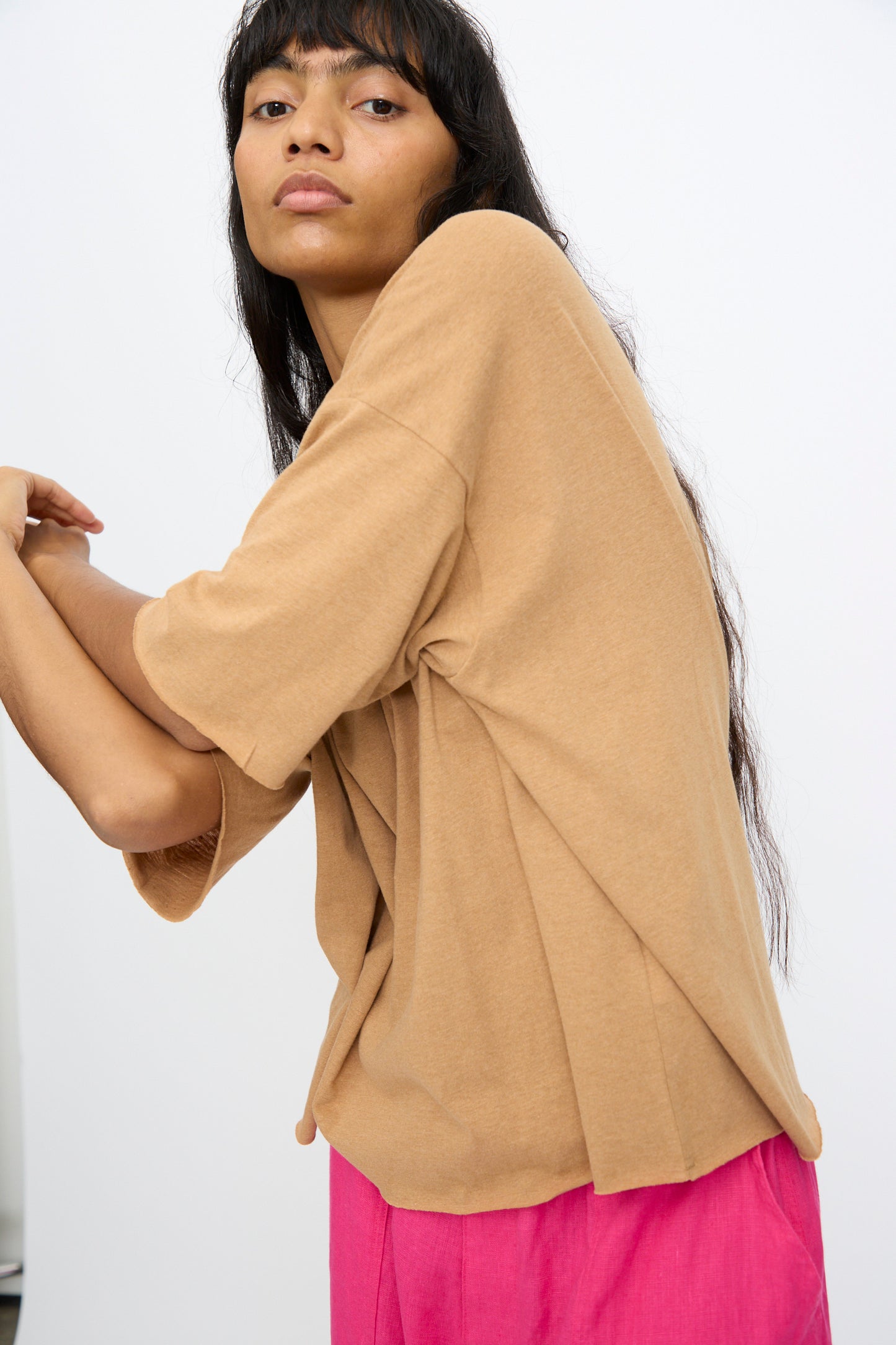 A woman with long dark hair models the Ichi Antiquités Cotton Pullover Tee in Camel, styled with bright pink pants, posing against a plain white background.