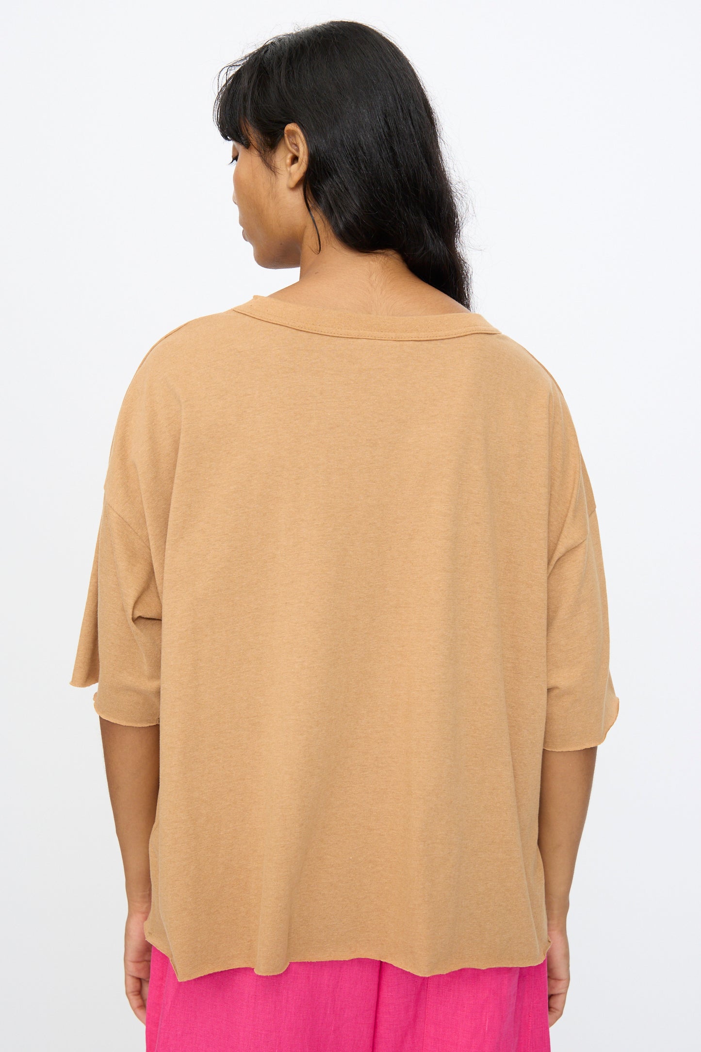 A person with long dark hair wears the Ichi Antiquités Cotton Pullover Tee in Camel, styled loosely and boxy, paired with bright pink pants, facing away from the camera against a plain light background.