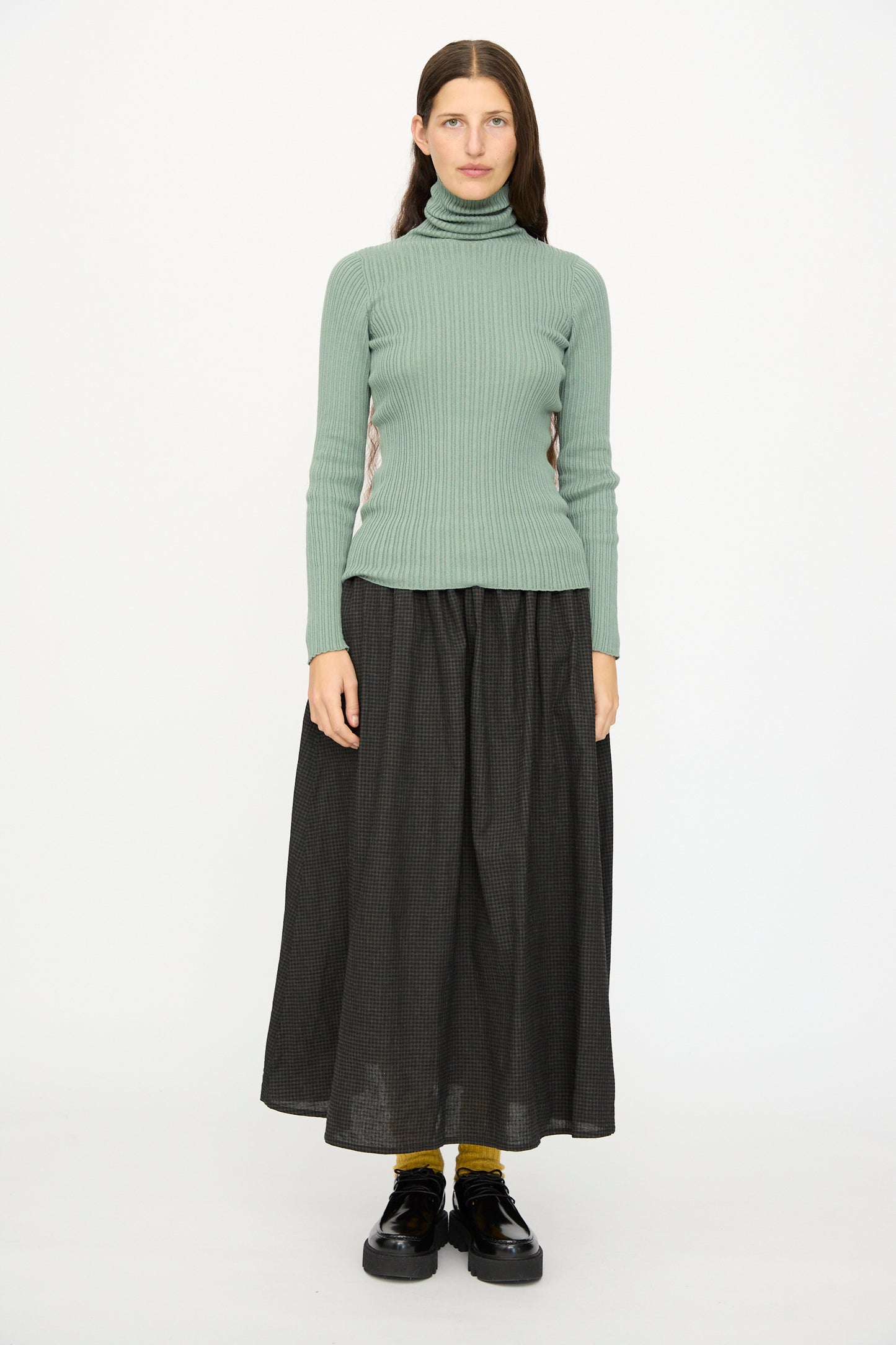A person faces forward against a white background, wearing the Ichi Antiquités Cotton Rib Knit Turtleneck in Green, a long dark skirt, yellow socks, and black shoes.