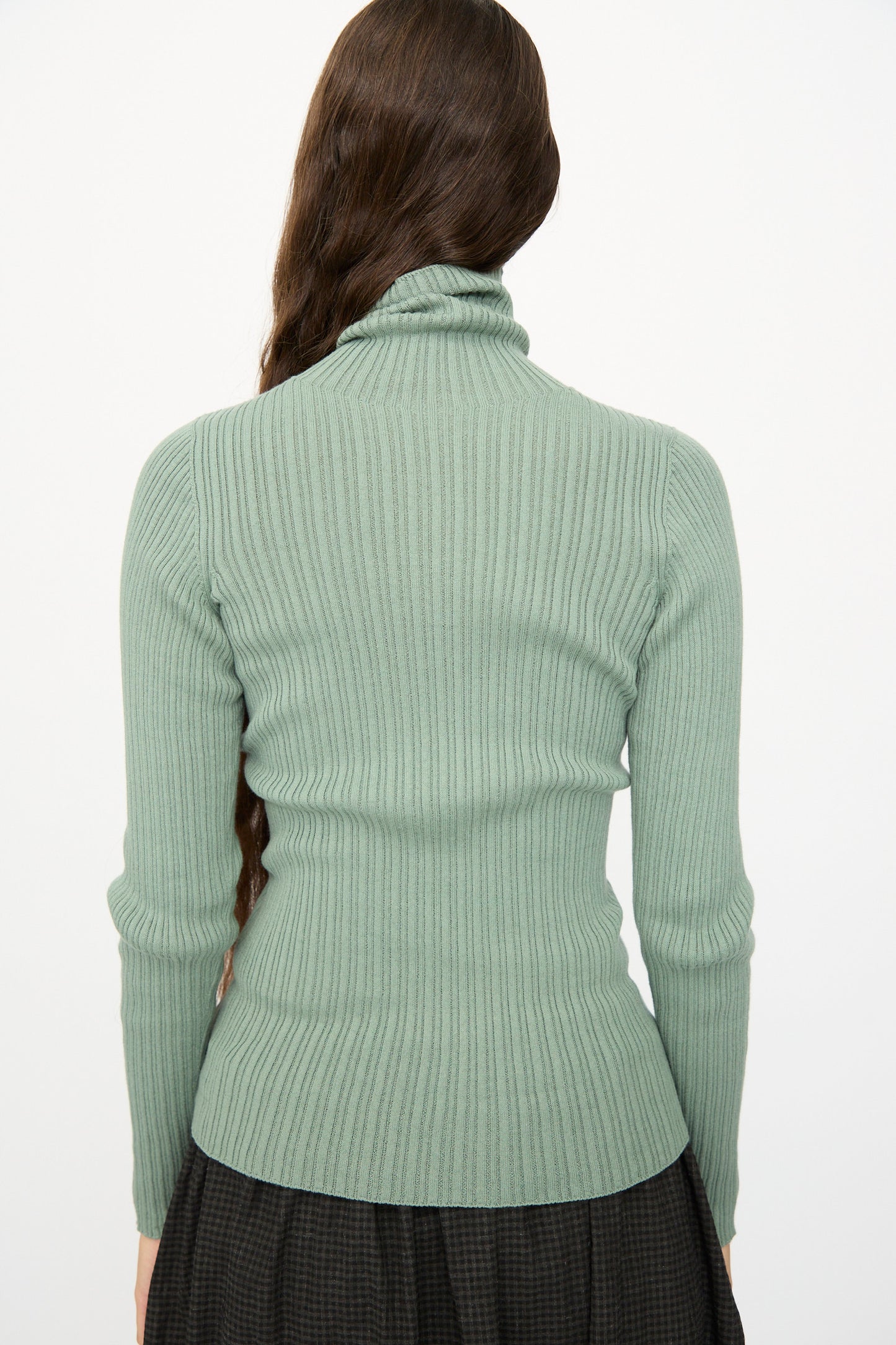A person with long brown hair, shown from the back against a white background, is wearing the Ichi Antiquités Cotton Rib Knit Turtleneck in Green and a dark skirt.