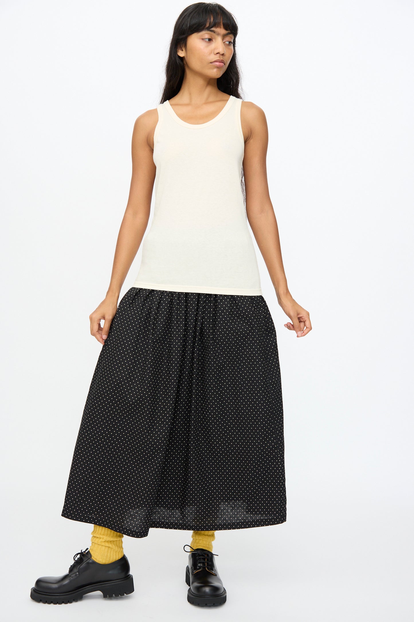 A woman wears the Ichi Antiquités Cotton Tank Top in Natural, styled with a long black skirt with white dots, yellow tights, and black lace-up shoes against a plain white background.