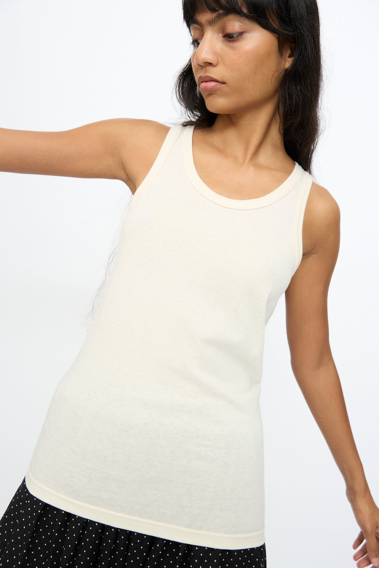 A person wears the Ichi Antiquités Cotton Tank Top in Natural, styled with a black skirt featuring white polka dots, standing against a plain, light background.