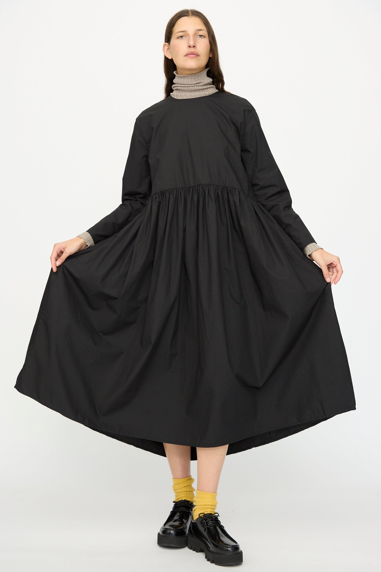 A person wearing the Ichi Antiquités Cotton Typewriter Dress in Black, layered over a gray turtleneck with yellow socks and black shoes, stands against a plain white background.