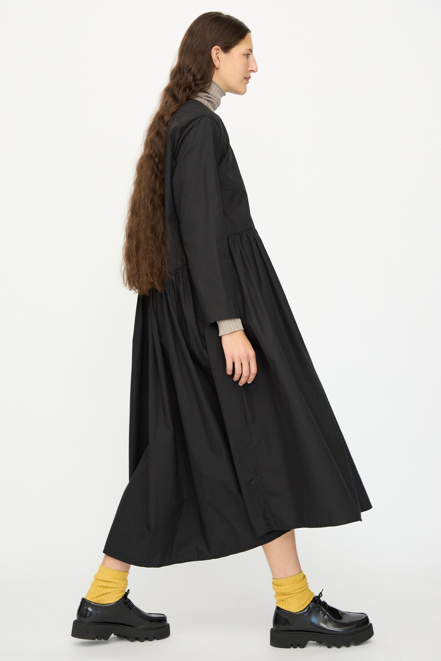 A person stands sideways against a plain white background wearing the Ichi Antiquités Cotton Typewriter Dress in Black, layered over a gray turtleneck, with black platform shoes and yellow socks.
