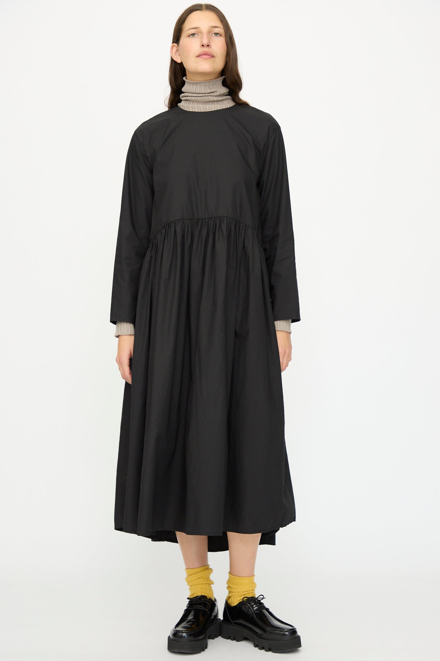 A person faces forward wearing the Ichi Antiquités Cotton Typewriter Dress in Black over a beige turtleneck, yellow socks, and black shoes, set against a plain white background.