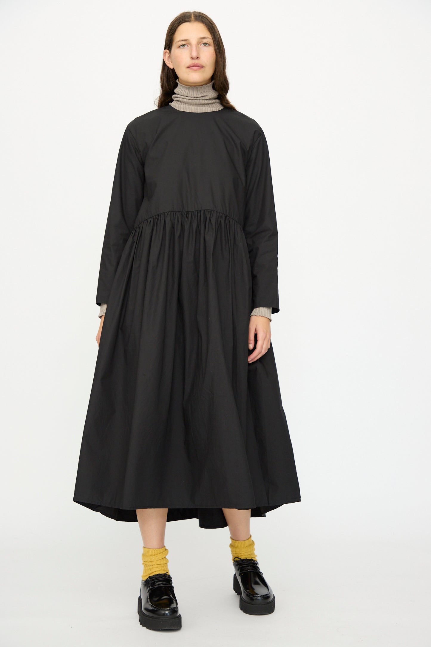 A person wears the Ichi Antiquités Cotton Typewriter Dress in Black over a beige turtleneck, styled with yellow socks and black shoes, standing against a plain background.