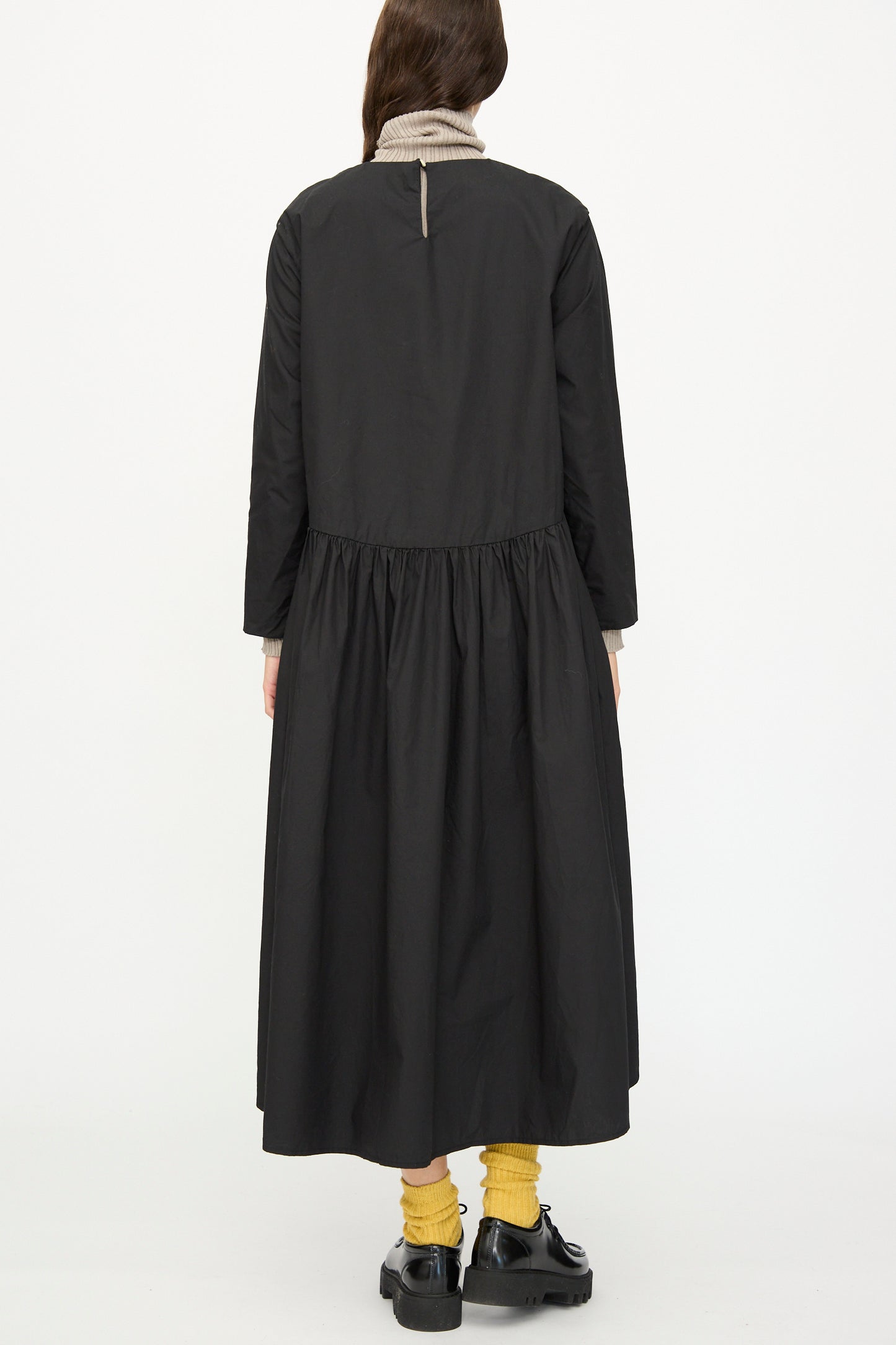 A person faces away, wearing the Ichi Antiquités Cotton Typewriter Dress in Black over a beige turtleneck, paired with yellow socks and black shoes, set against a plain white background.