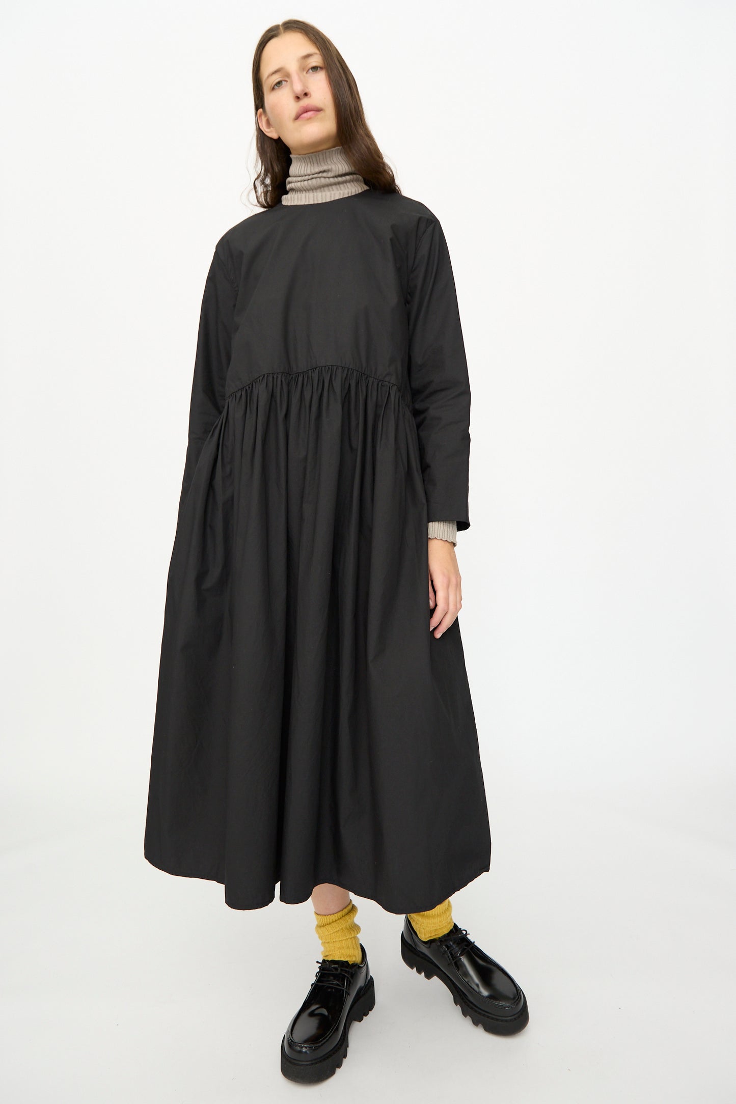 A person models the Ichi Antiquités Cotton Typewriter Dress in Black over a beige turtleneck, paired with yellow socks and black shoes, against a plain white background.