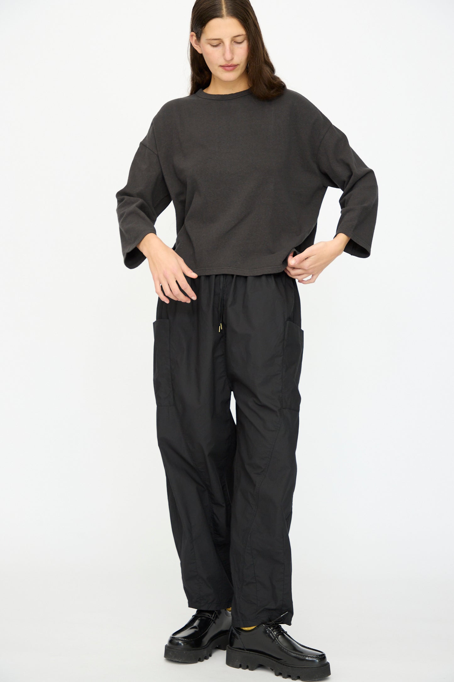 A person stands against a white background wearing a loose dark gray long-sleeve top, Ichi Antiquités Cotton Typewriter Pants in Black, and black platform shoes.