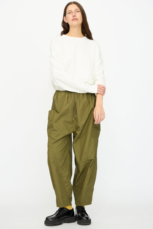 A person stands against a plain white background wearing a long-sleeve white top, Ichi Antiquités Cotton Typewriter Pants in Khaki, and black shoes.