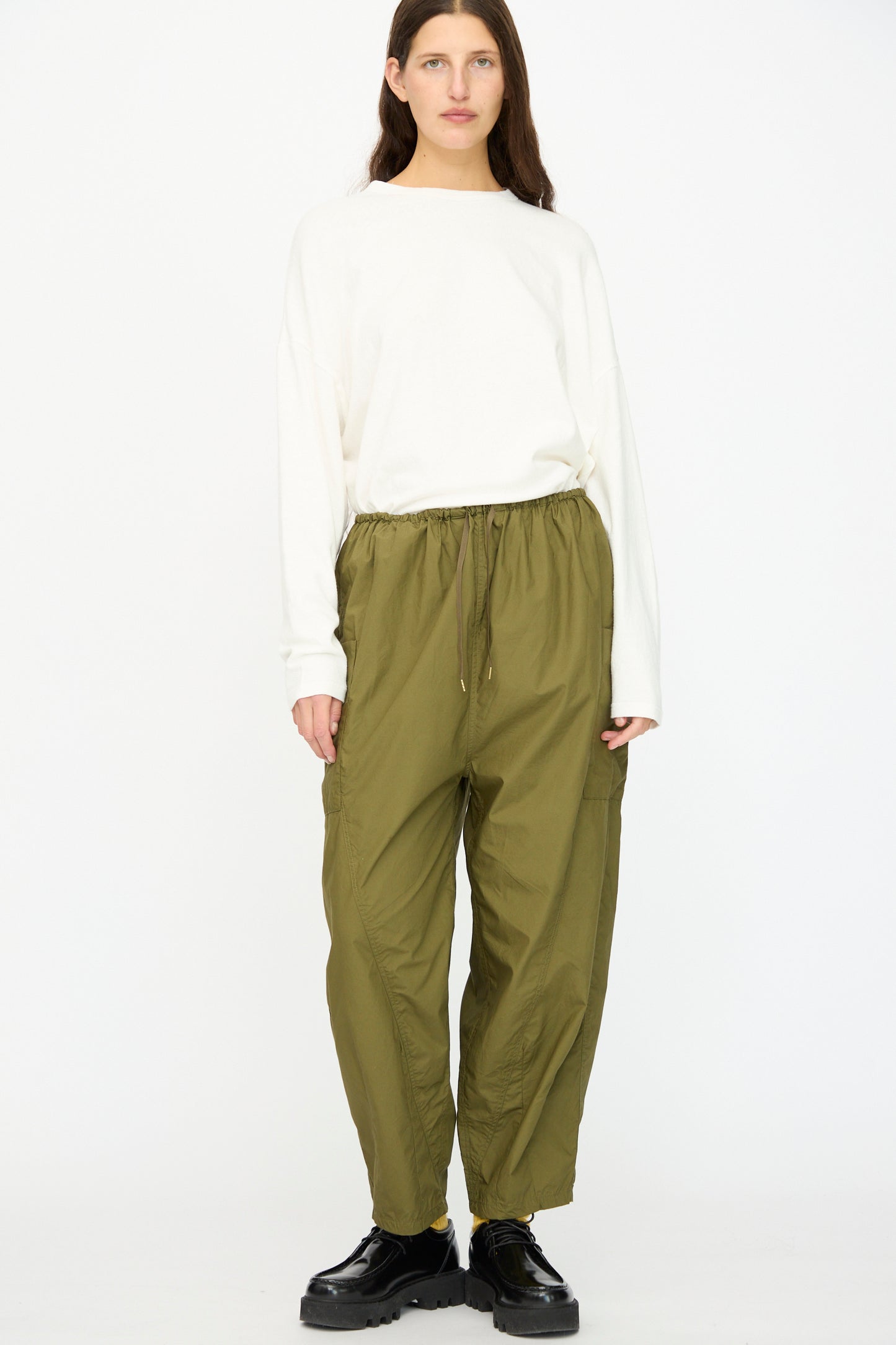 A person stands facing forward wearing a loose white long-sleeve shirt, Ichi Antiquités Cotton Typewriter Pants in Khaki with a drawstring, and black shoes against a plain white background.
