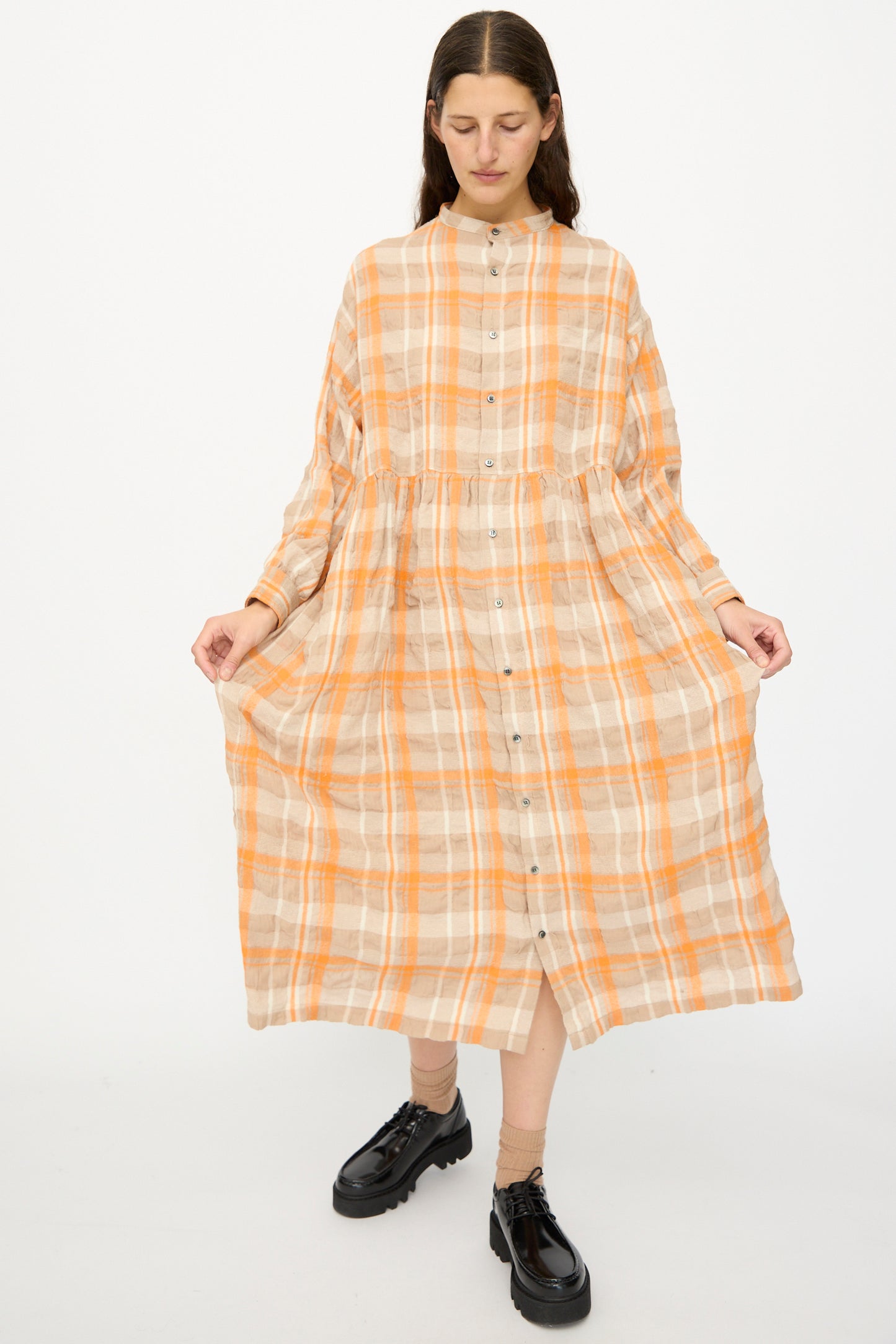 A person stands against a white background wearing the Ichi Antiquités Cotton Wool Tartan Check Dress in orange, featuring long sleeves, an oversized silhouette, and front buttons, paired with black lace-up shoes.