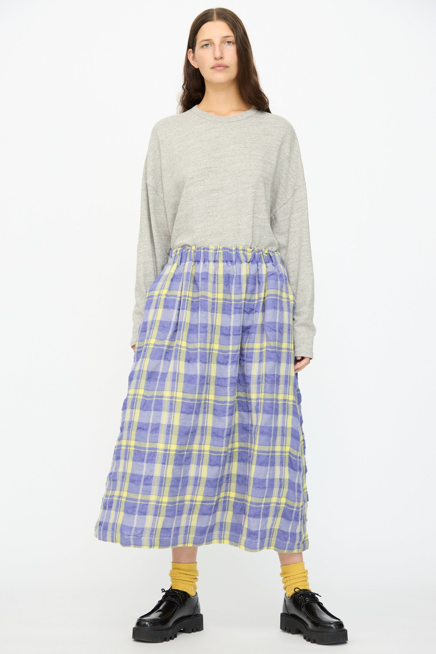 A person stands facing forward against a white background, wearing a gray long-sleeve top, Ichi Antiquités’ Cotton Wool Tartan Check Skirt in Blue with an elastic waist, yellow socks, and black platform shoes.