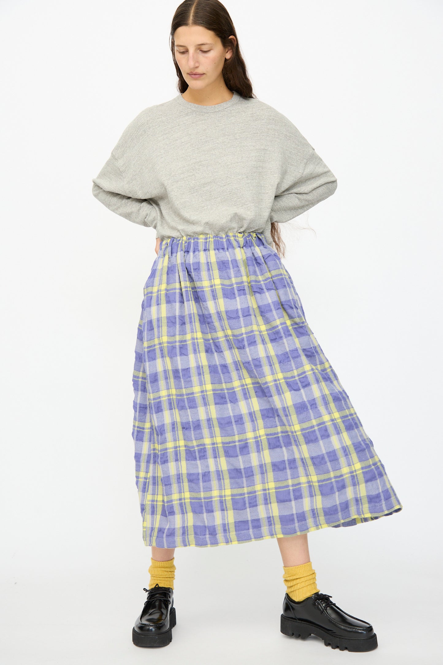 A person stands with hands on hips, wearing a loose grey sweater, yellow socks, black shoes, and the Ichi Antiquités Cotton Wool Tartan Check Skirt in Blue against a plain white background.