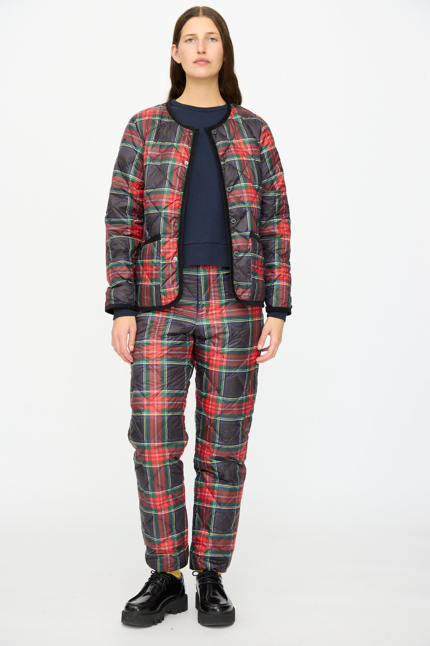 A person faces forward against a plain white background, wearing the Ichi Antiquités Down Jacket in Black Tartan over a navy top, paired with matching pants featuring black corduroy trim and black shoes.