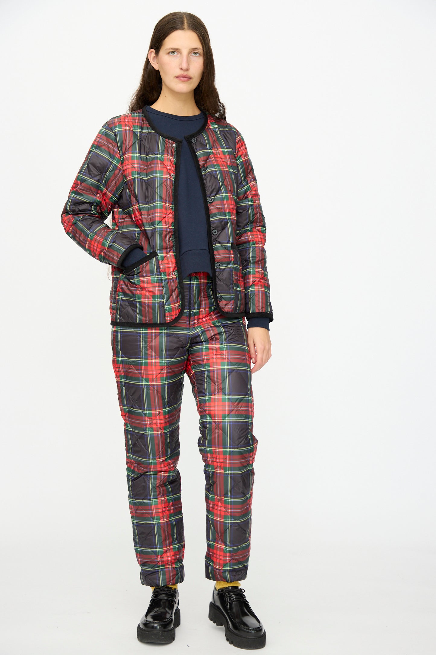 A person stands against a white background wearing the Ichi Antiquités Down Jacket in Black Tartan, styled with matching red and green plaid pants, a black shirt, and black shoes.