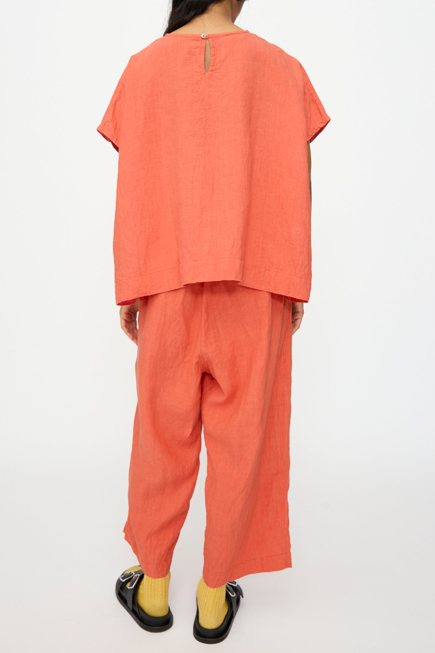 A person stands with their back to the camera in a boxy fit French Linen Pullover in Orange by Ichi Antiquités, paired with pants, yellow socks, and black sandals against a plain background. This relaxed outfit is made in Japan.