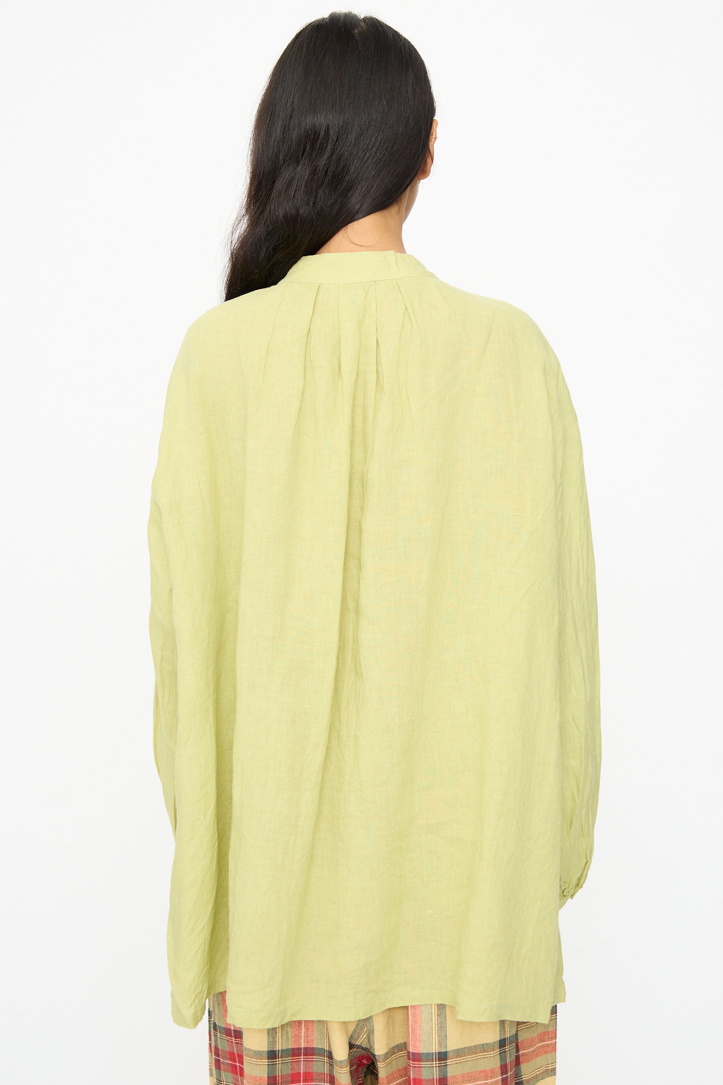 A person with long dark hair is shown from the back, wearing the Ichi Antiquités French Linen Shirt in Pistachio, featuring a loose fit and band collar, paired with plaid pants against a plain white background.