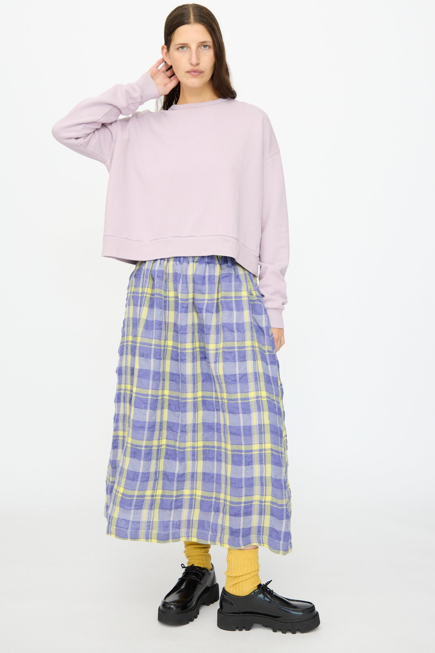 A person stands against a white background wearing the Ichi Antiquités French Terry Cotton Pullover in Lavender, paired with a blue and yellow plaid skirt, yellow socks, and black platform shoes.