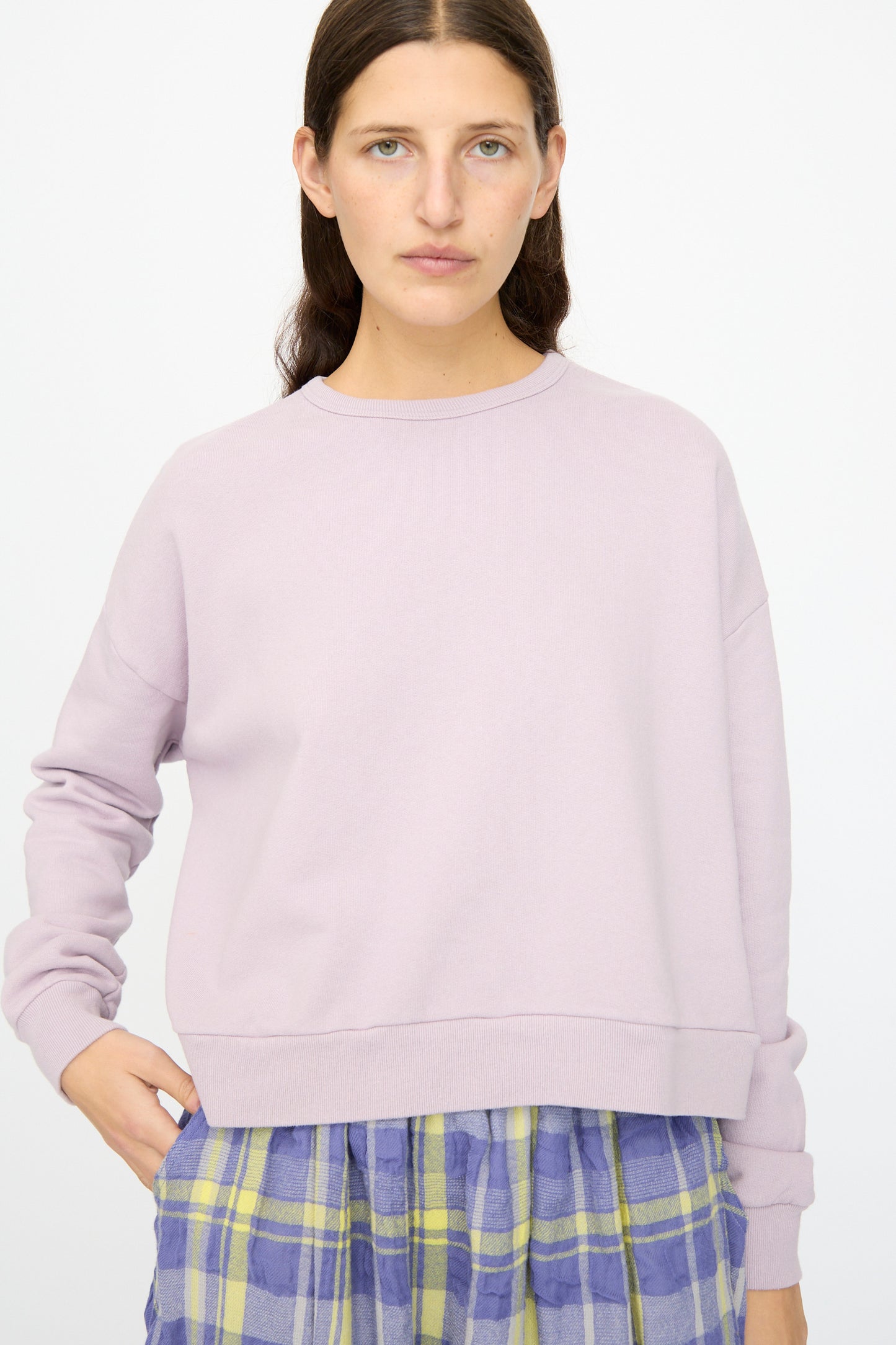 A person with long brown hair models the Ichi Antiquités French Terry Cotton Pullover in Lavender, paired with a blue and yellow plaid skirt, against a plain white background.