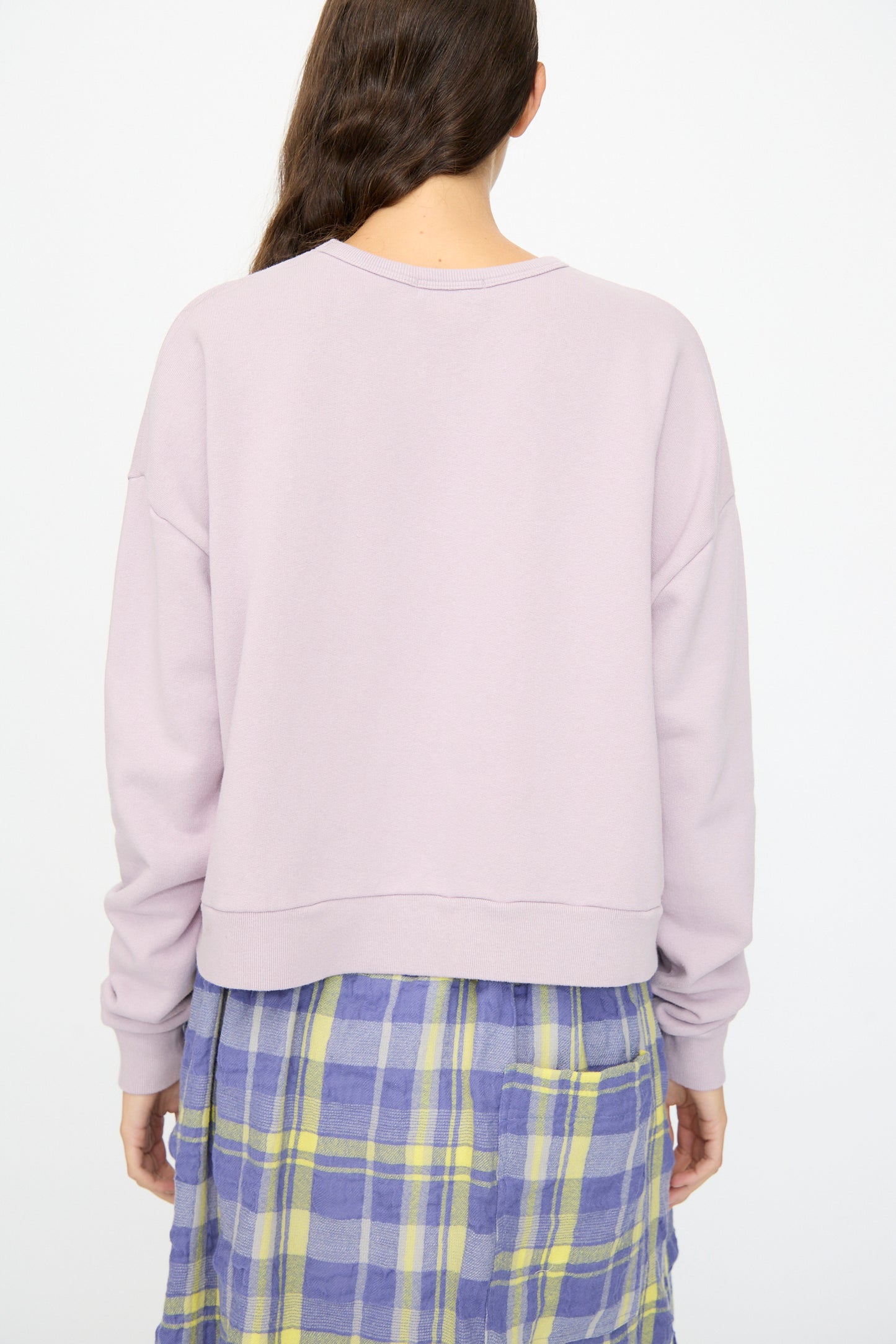 A person with long brown hair is shown from the back, wearing the Ichi Antiquités French Terry Cotton Pullover in Lavender and a blue and yellow plaid skirt.