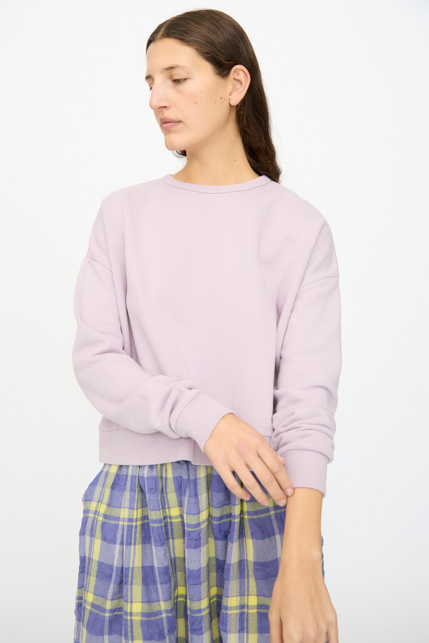A person with long brown hair models the Ichi Antiquités French Terry Cotton Pullover in Lavender, paired with a blue and yellow plaid skirt, standing against a plain white background.