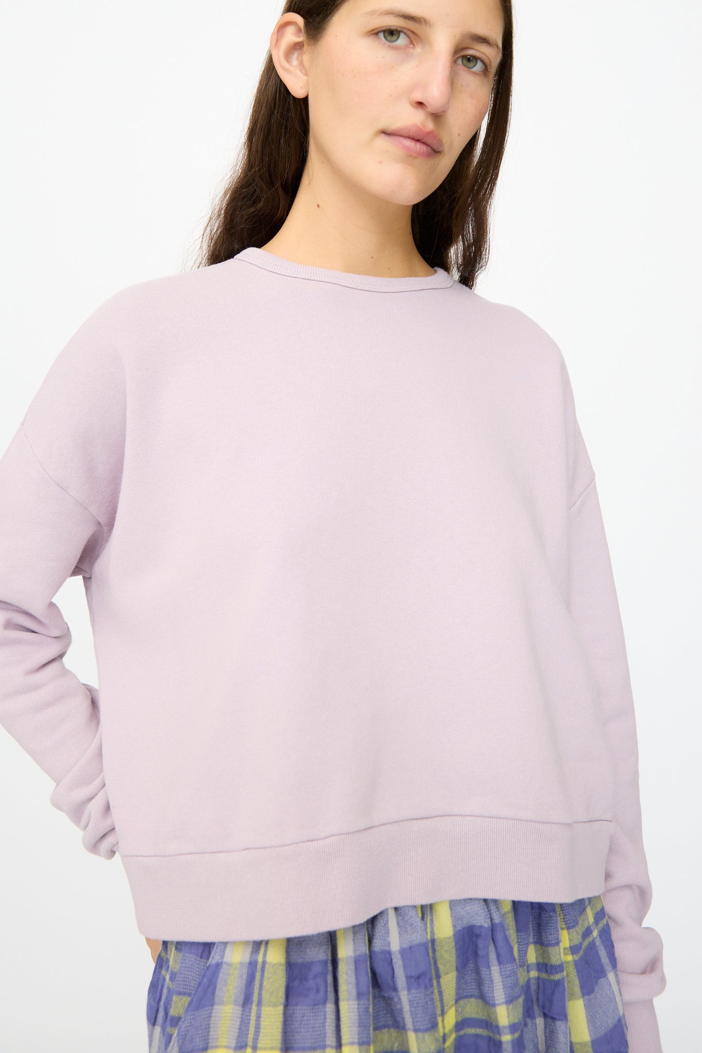 A person stands against a plain background wearing the Ichi Antiquités French Terry Cotton Pullover in Lavender and a plaid skirt with blue and yellow tones.