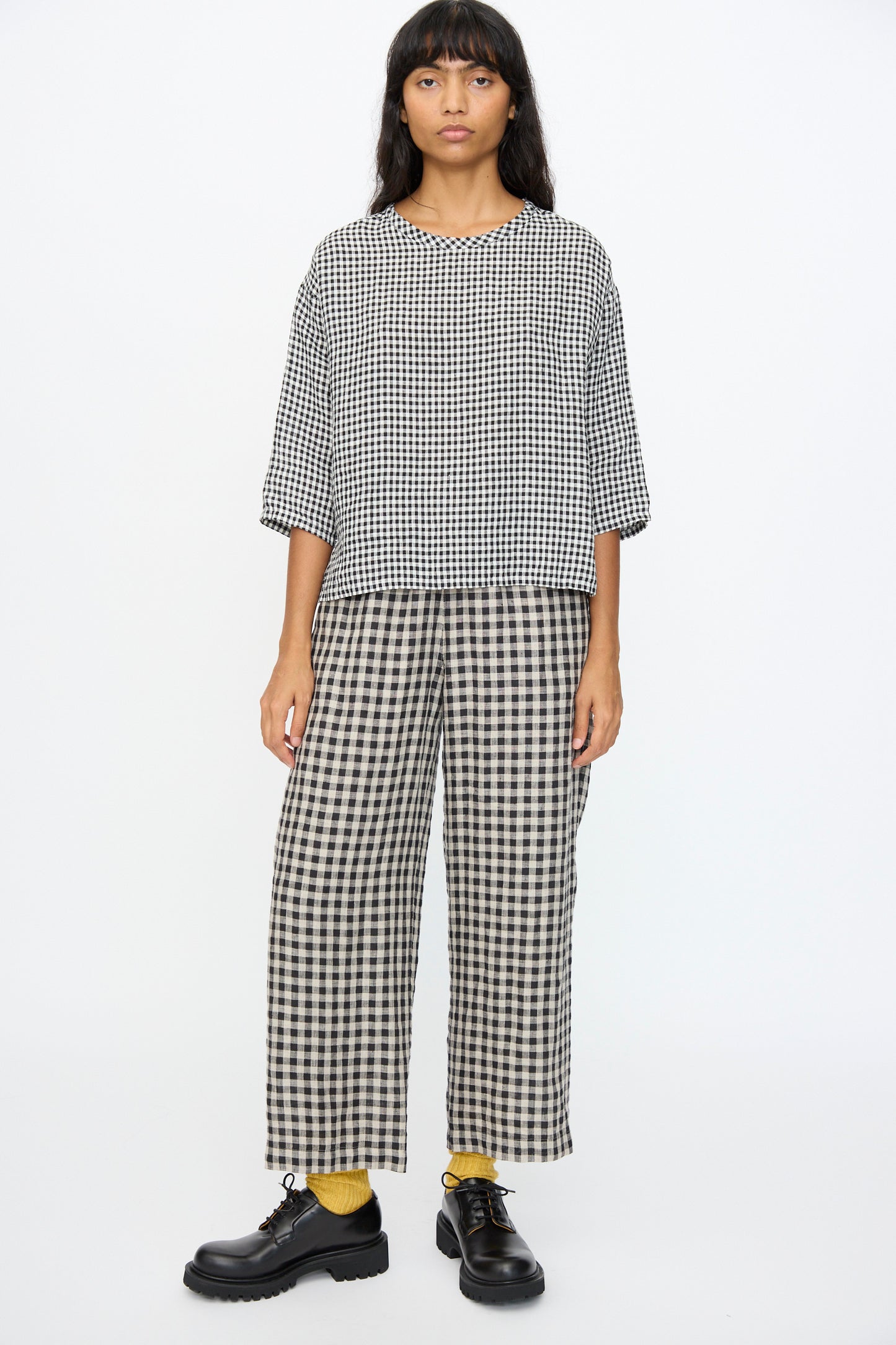 A person stands against a plain background wearing the Ichi Antiquités Gingham Check Blouse in White and Black, featuring an oversized fit and crafted in Japan, paired with matching wide-leg pants, black shoes, and yellow socks.