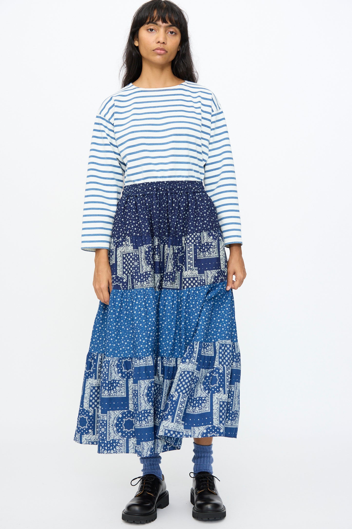A woman stands against a plain background wearing a white and blue striped long-sleeve shirt, black shoes, and the Ichi Antiquités Indigo Flower Skirt in Navy Mix.