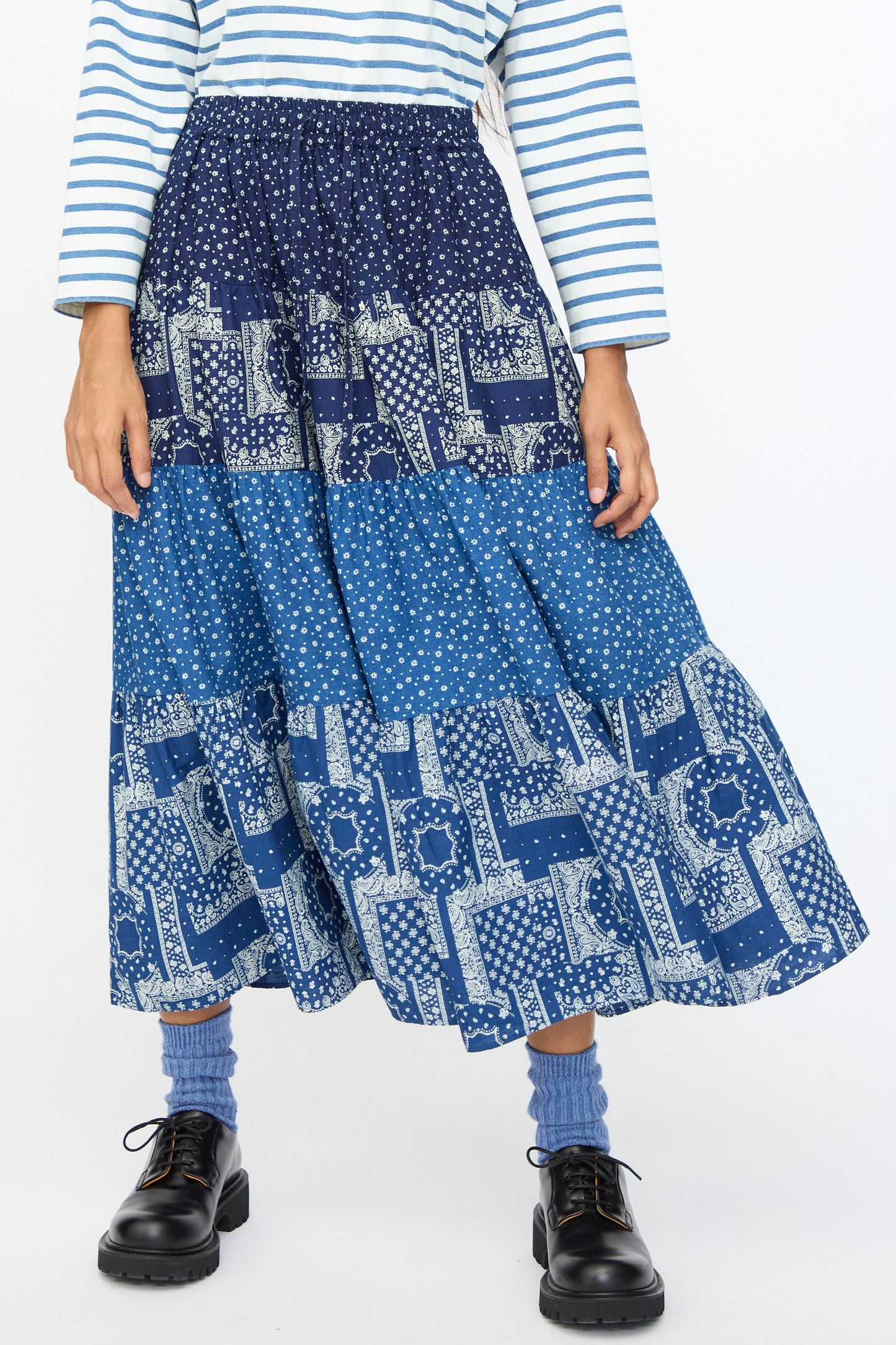 A person wears the Ichi Antiquités Indigo Flower Skirt in Navy Mix, styled with a blue and white striped long-sleeve shirt, blue socks, and black shoes, standing against a plain background.