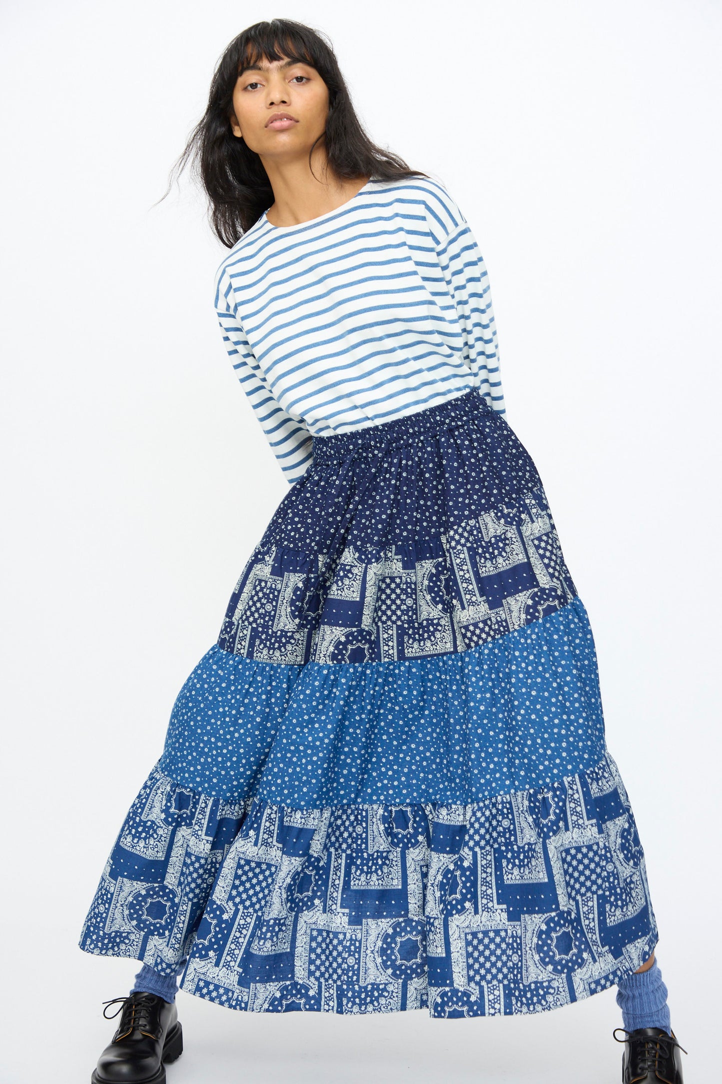 A woman wears the Indigo Flower Skirt in Navy Mix by Ichi Antiquités, paired with a blue and white striped long-sleeve shirt and black boots, standing against a plain white background.