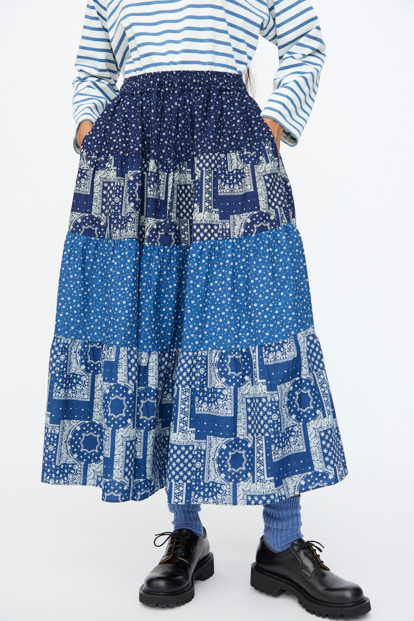 A person wearing a striped long-sleeve shirt, blue socks, black shoes, and the Indigo Flower Skirt in Navy Mix by Ichi Antiquités stands with hands in skirt pockets.