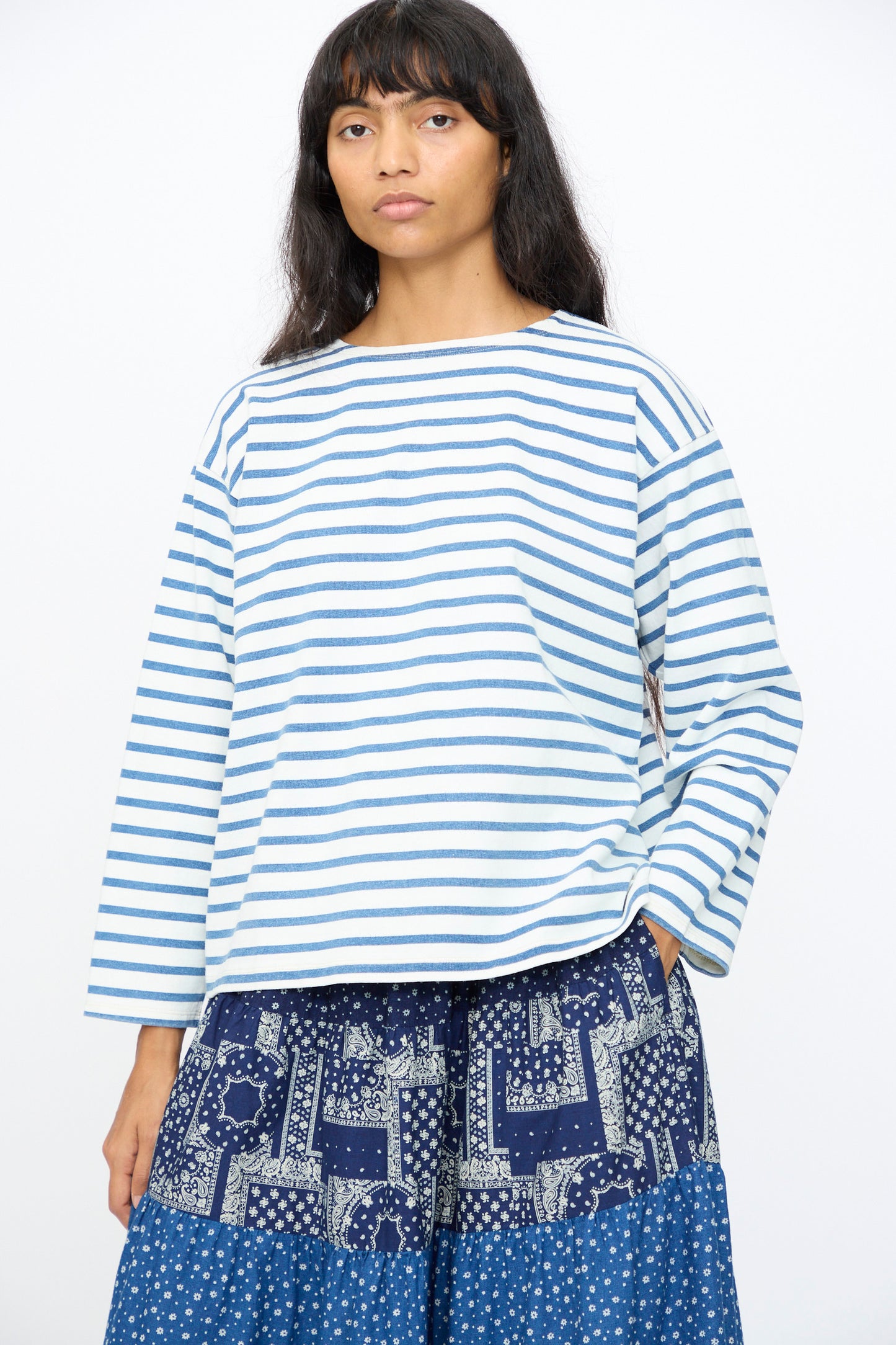 A woman wears the Ichi Antiquités Indigo Pullover Tee in White and Blue, paired with a blue patterned skirt, standing against a plain white background.