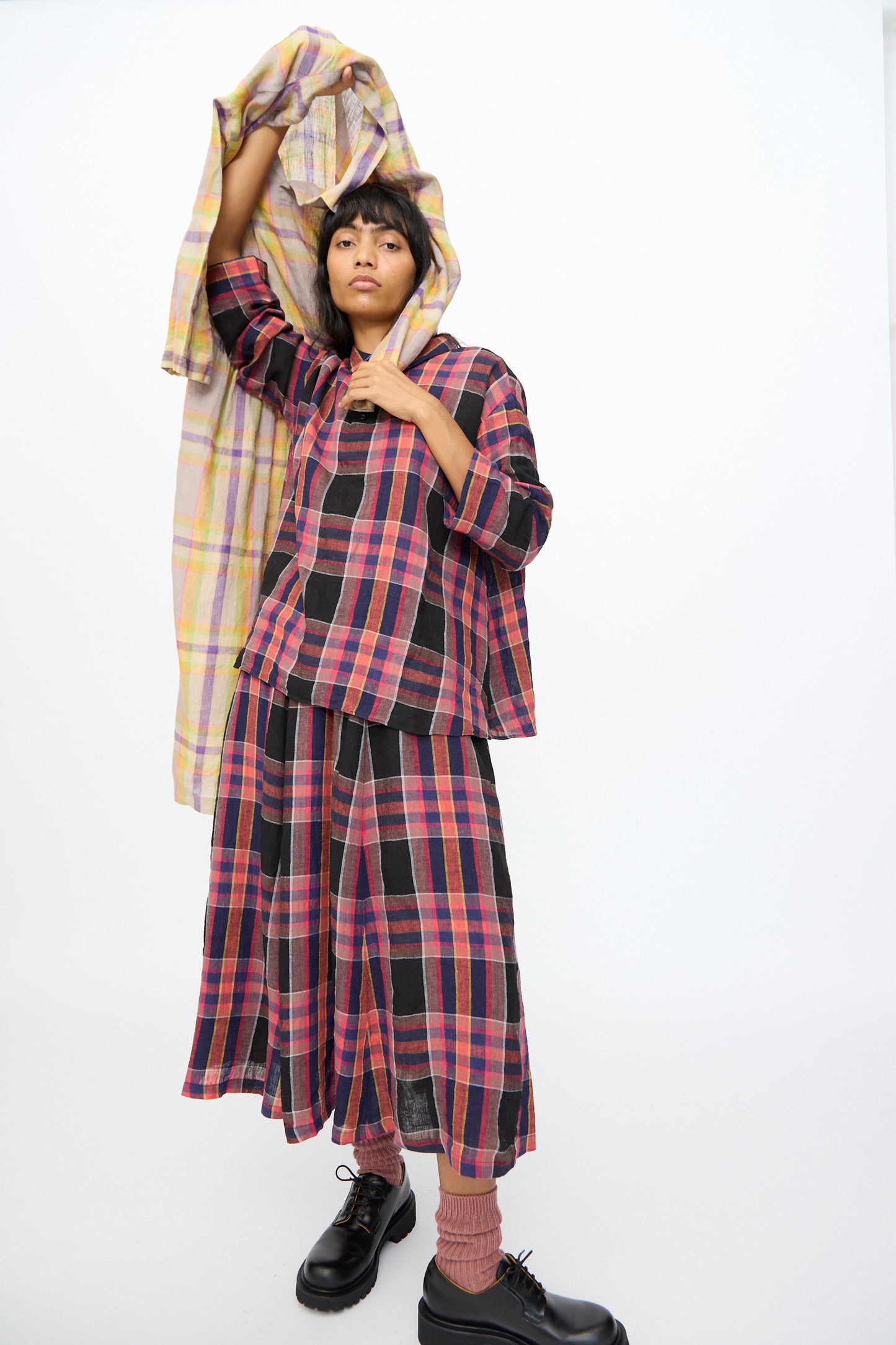 Model wearing the Ichi Antiquités Linen Check Skirt in Black, lifting a matching plaid fabric overhead, set against a plain white background.