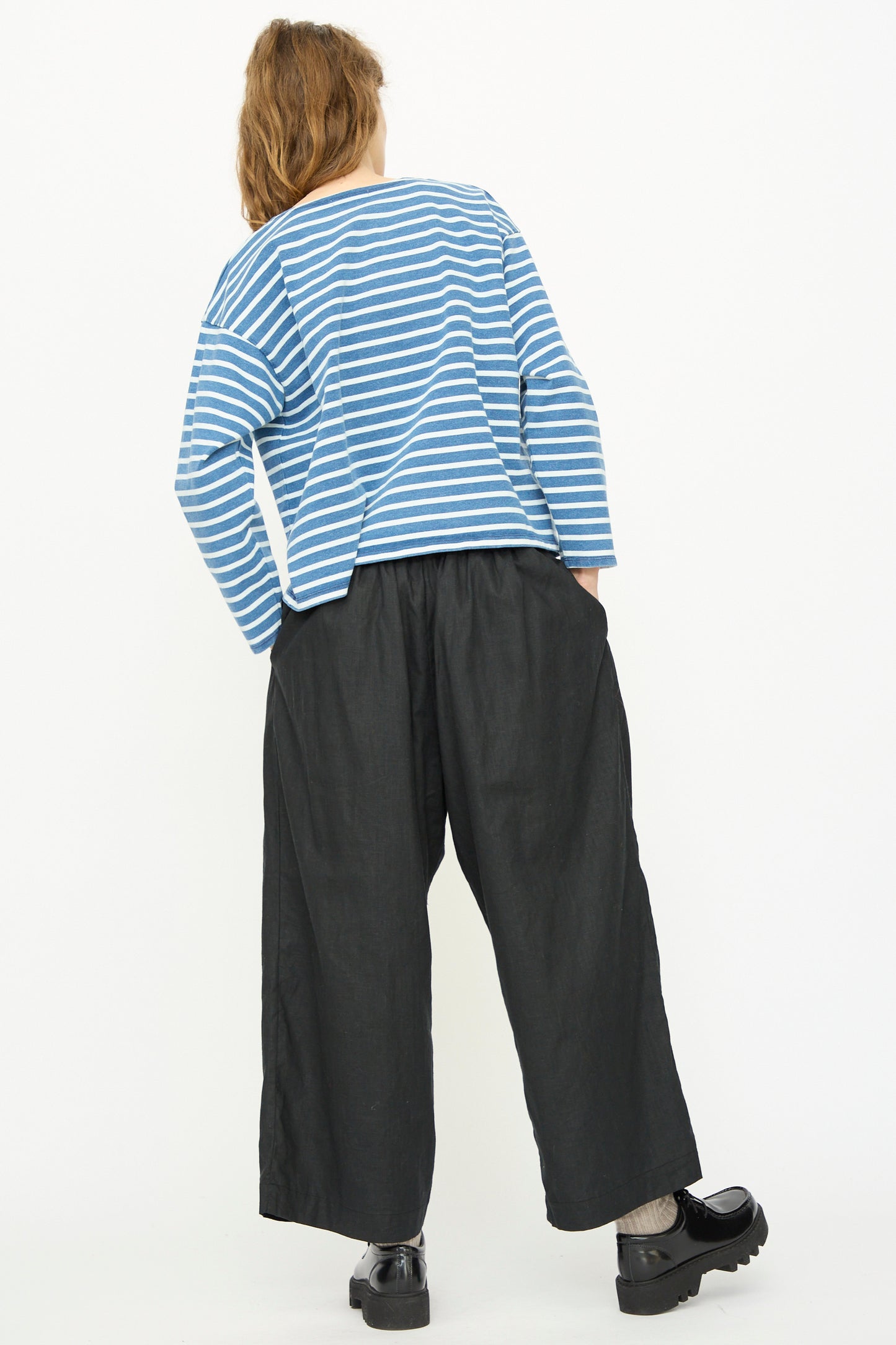 Person facing away from the camera wears a blue and white striped long-sleeve shirt, Ichi Antiquités Linen Cotton Canvas Pant in Black with an elastic waist, and black shoes against a plain white background.